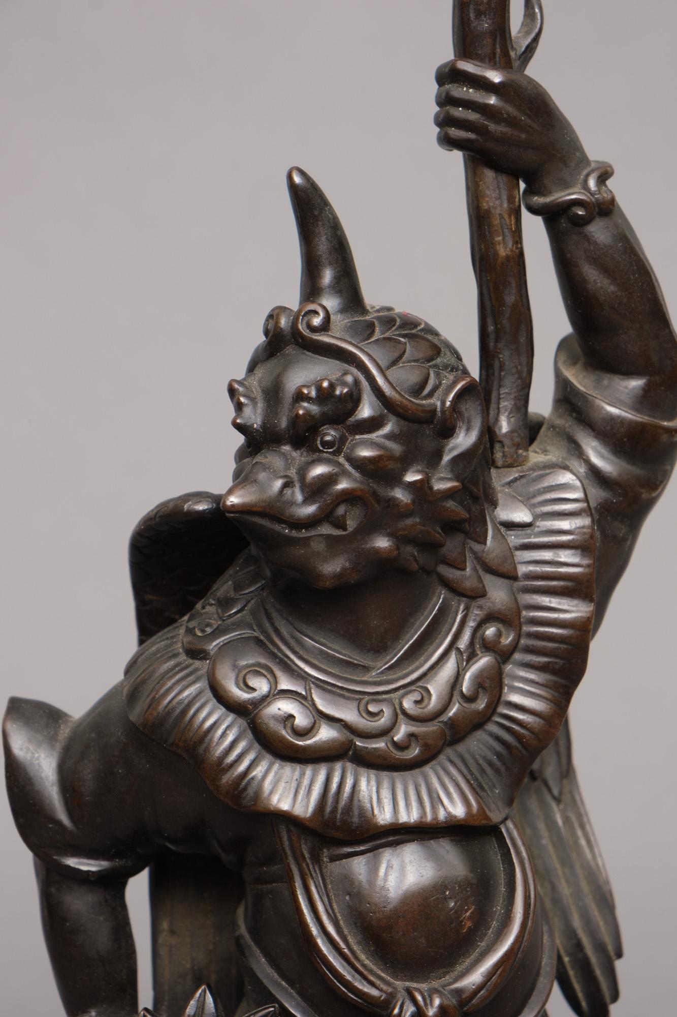 Very refined Japanese bronze okimono of a ‘karasu tengu’ holding a candlestick.

Well cast as a tengu in half-human, half-bird form standing atop a pile of leaves on a rounded square base with lappet and wave diaper to the sides and supported on
