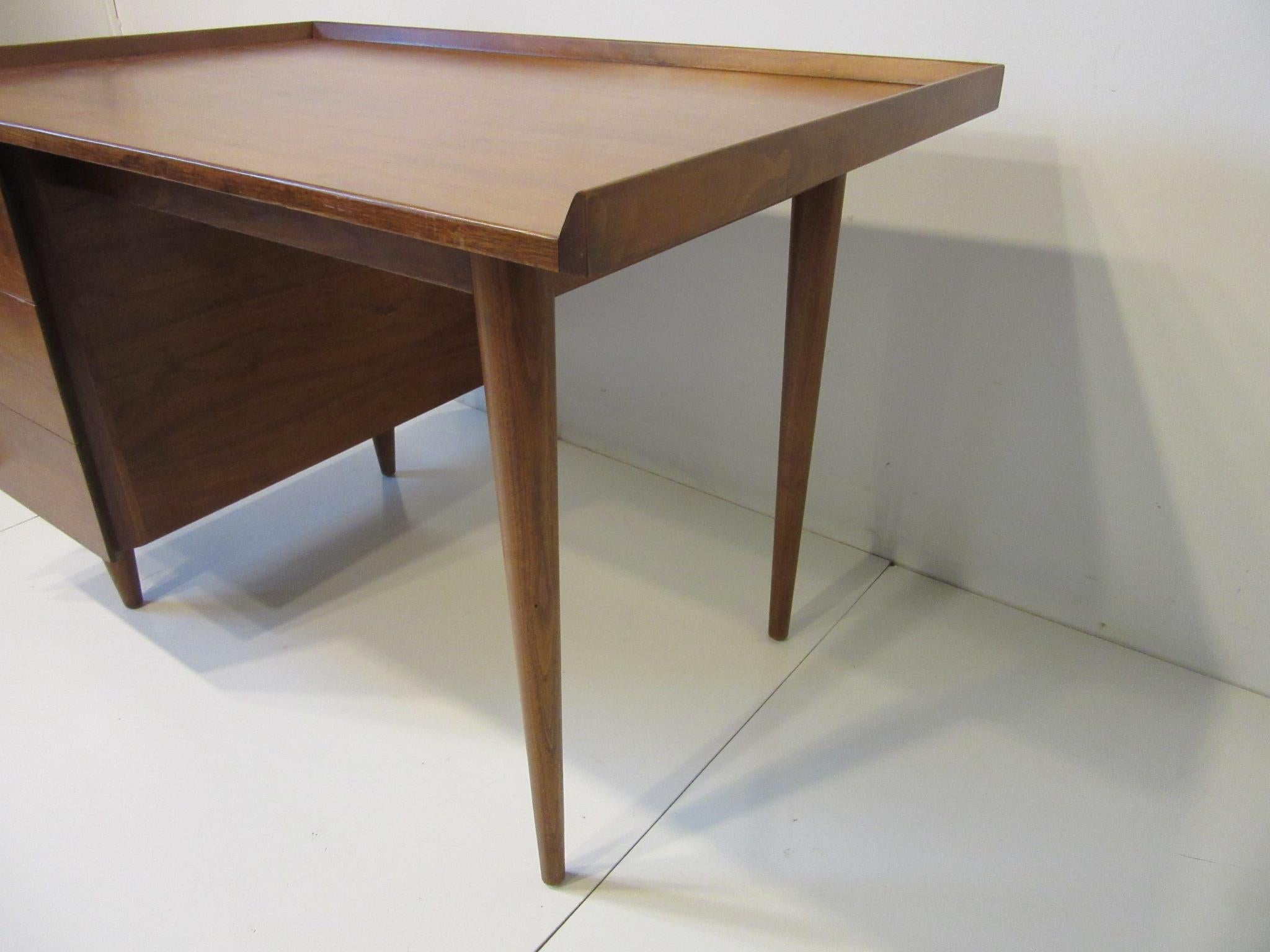 A smaller sized dark walnut single pedestal desk with three angled side drawers , and raised edges to the sides and back of the top having a compartment and pen holder to the top drawer . A well crafted and designed piece from the iconic Mid Century