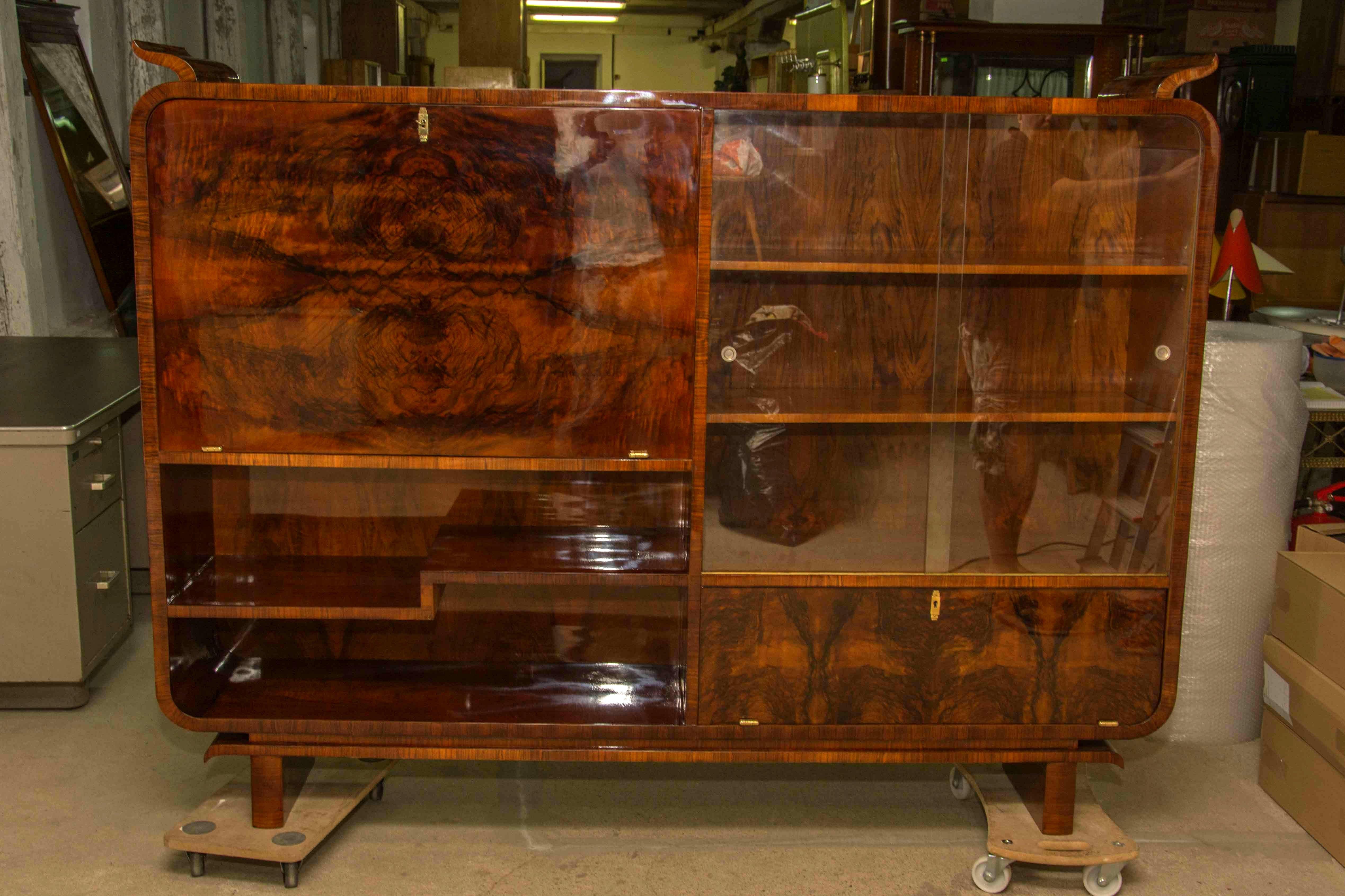 This walnut library cabinet / bookcase Art Deco was designed and produced in the 1930s in Bohemia. The upper part of unit consists of a glazed space on the left with a bar on the right. The lower part consists of a storage space, closed and opened.