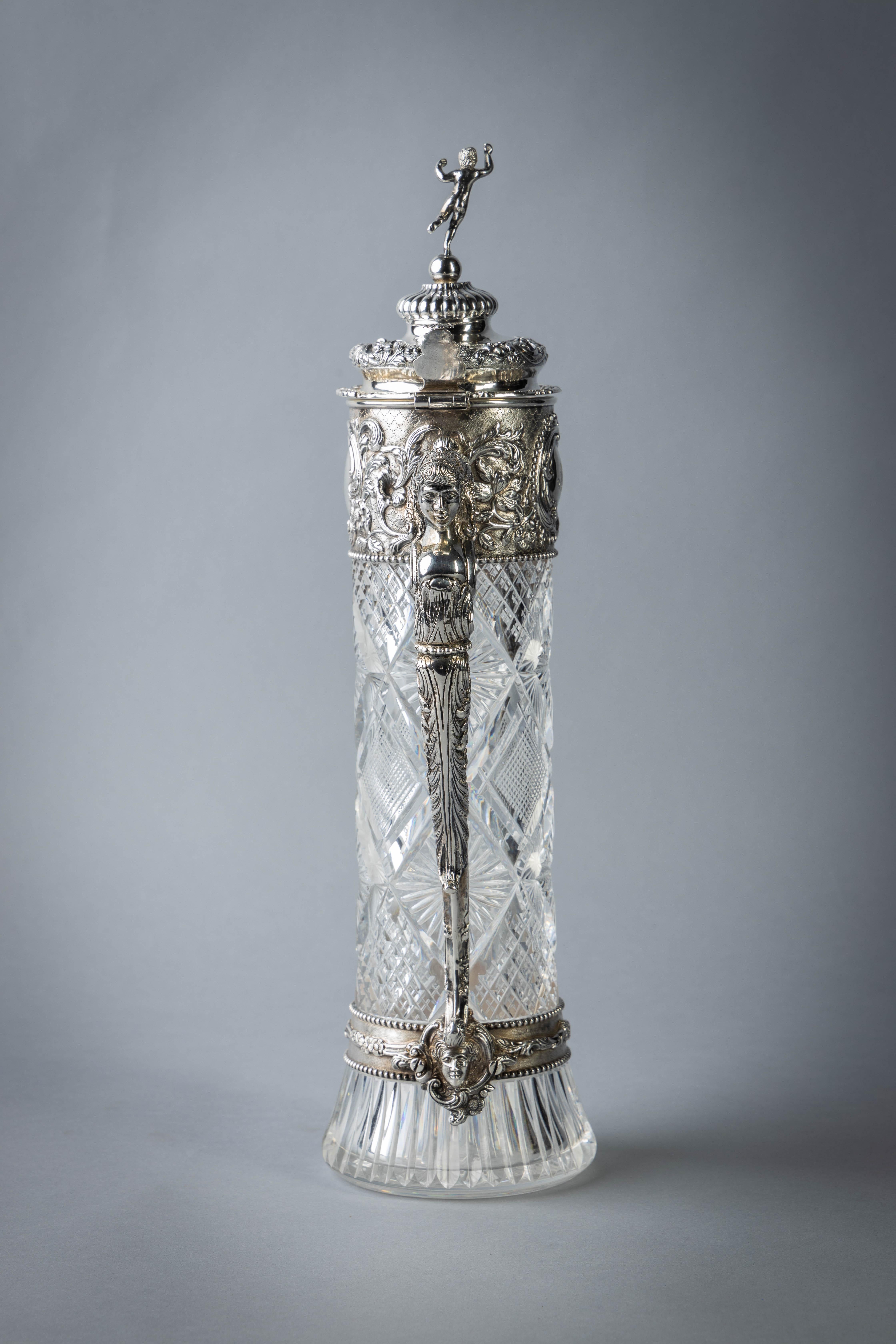 The glass body with vari-cut motifs, the silver with finely chased decoration with mask head spout and handle, the finial with putti standing on ball. Monogram within cartouche.