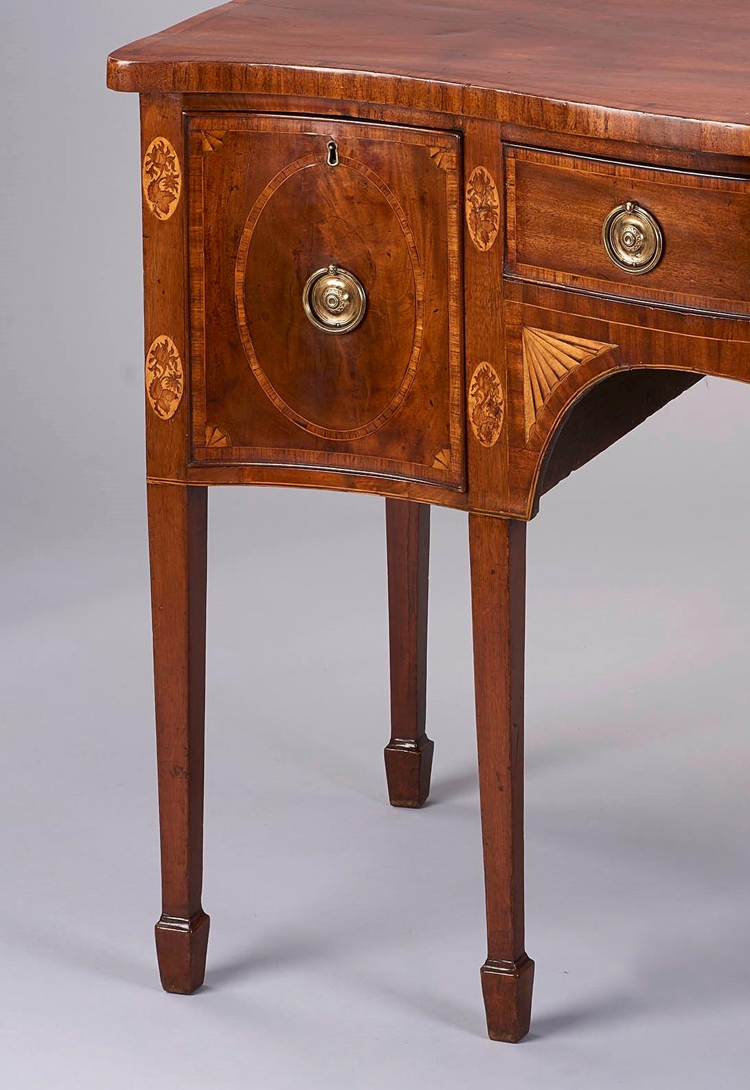 Very fine George III period mahogany serpentine-fronted sideboard in the Sheraton style; of particularly good color and patina; fitted with three cross-banded drawers, those on the left and right with inlaid oval panels; the stiles each inlaid with