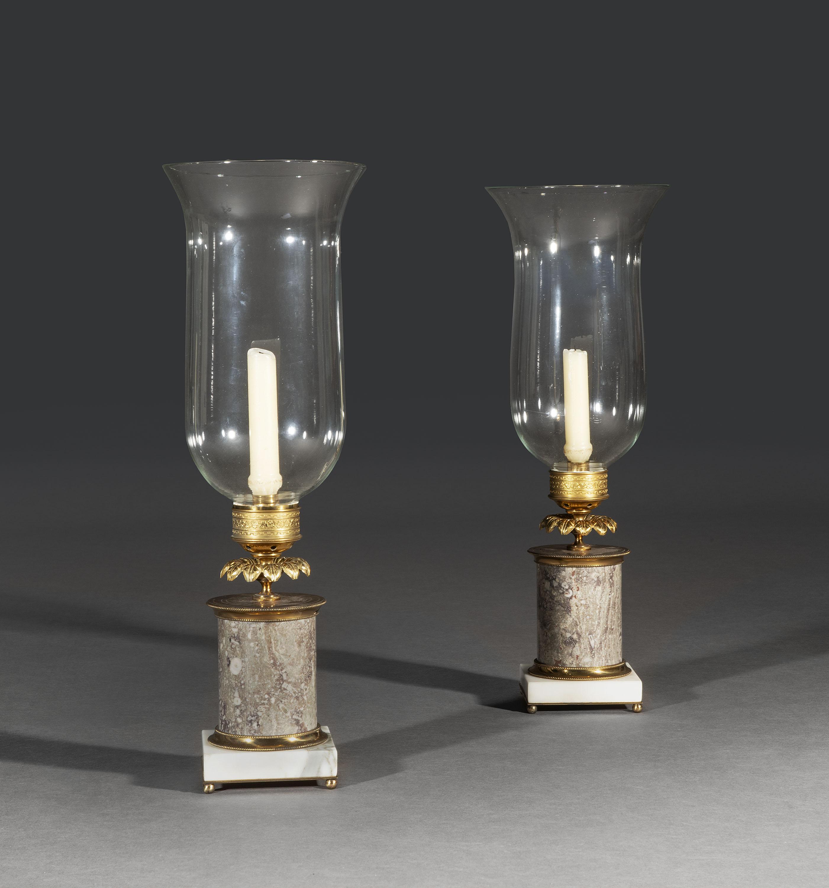 A rare pair of French ormolu and fossil marble table candlesticks with storm-shades with grey columns mounted on square white statuary marble bases with finely gilded mounts and ball feet, surmounted by leafy calyx and decorative sconces with their