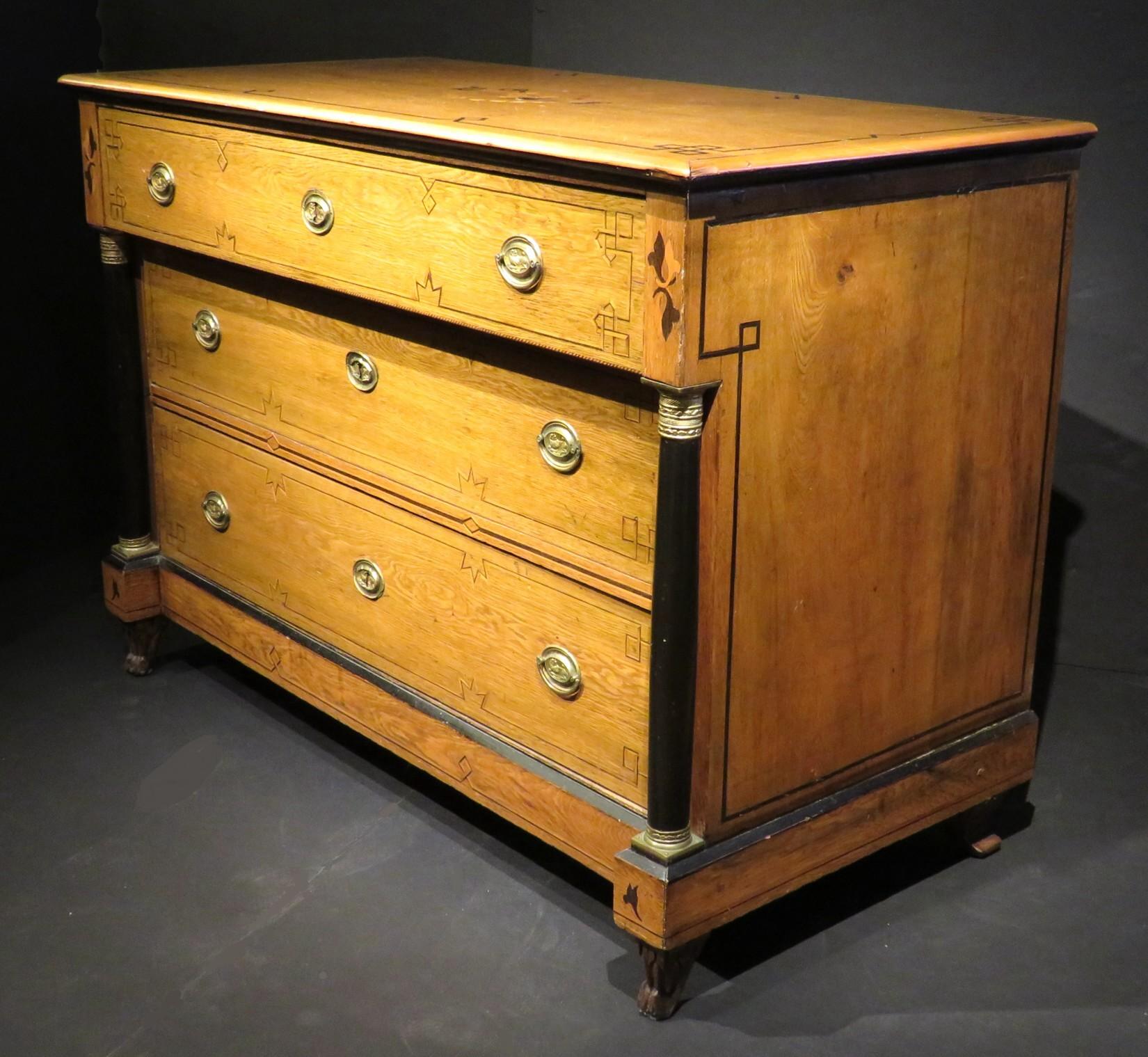 A very handsome & capacious three drawer commode in highly grained ash, the top inlaid with rectilinear parquetry motifs in German walnut, surrounding a heart-shaped marquetry motif centred by a silvered Christogram (IHS), intersected by marquetry
