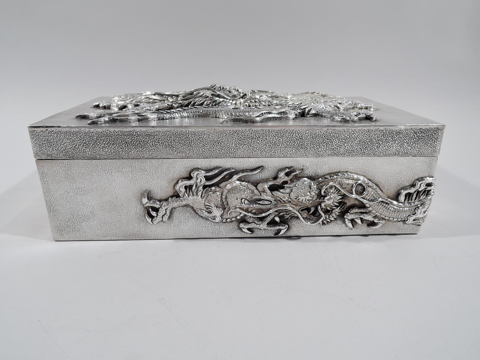 Large Japanese silver box, circa 1910. Rectangular with straight sides and hinged cover. all-over stippling. On cover is applied a dragon, all scales, fangs, and talons with slithering serpentine body. Same two wraparound sides. Fearsome protectors