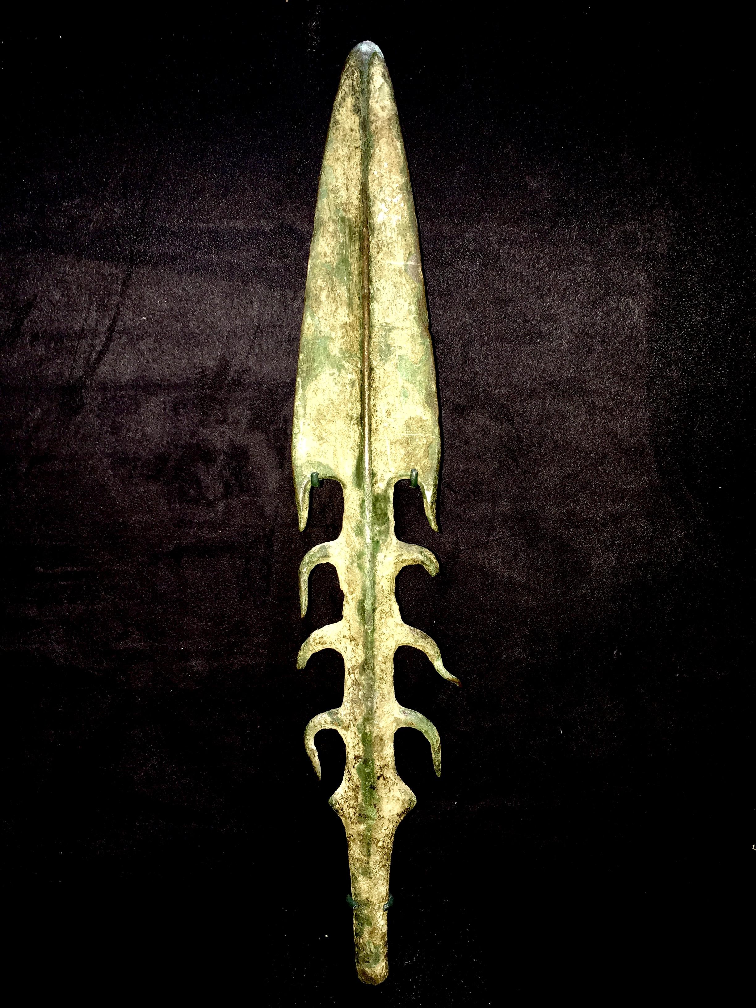 A substantial bronze spearhead or harpoon with thick midrib, triangular barbed blade, three pairs of lateral barbs and a pair of pierced spikes above a round-section tang. Very rare. This piece comes with a custom Stand in black matte finish and
