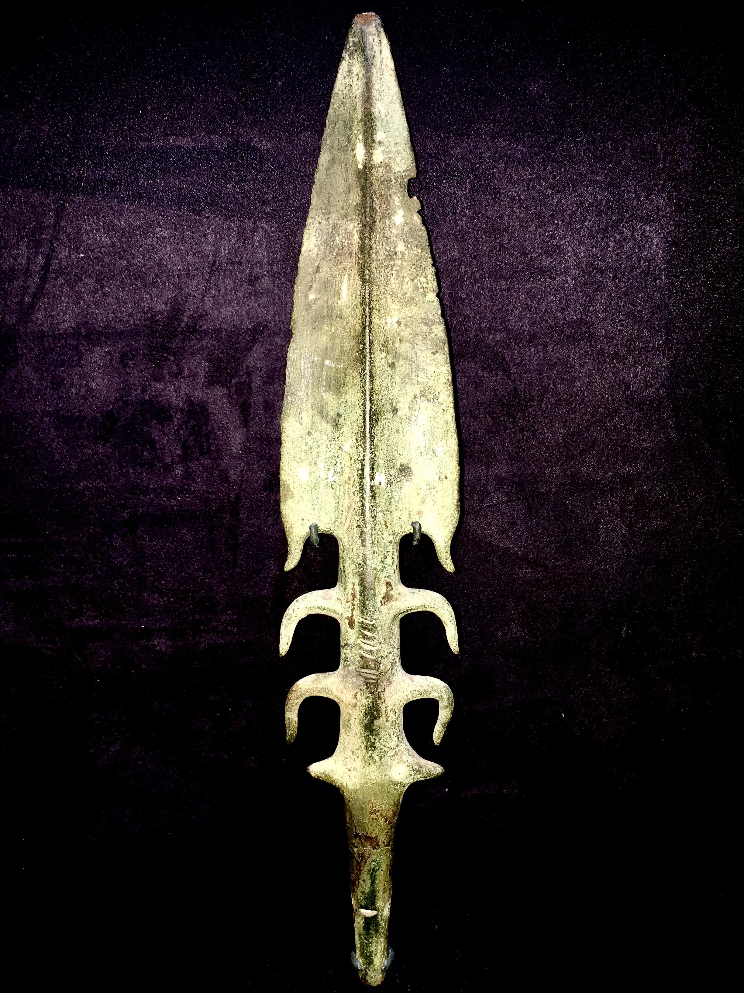 A substantial bronze spearhead or harpoon with thick midrib, triangular barbed blade, two pairs of lateral barbs and a pair of pierced spikes above a round-section tang. Very rare. This piece comes with a custom stand in black matte finish and