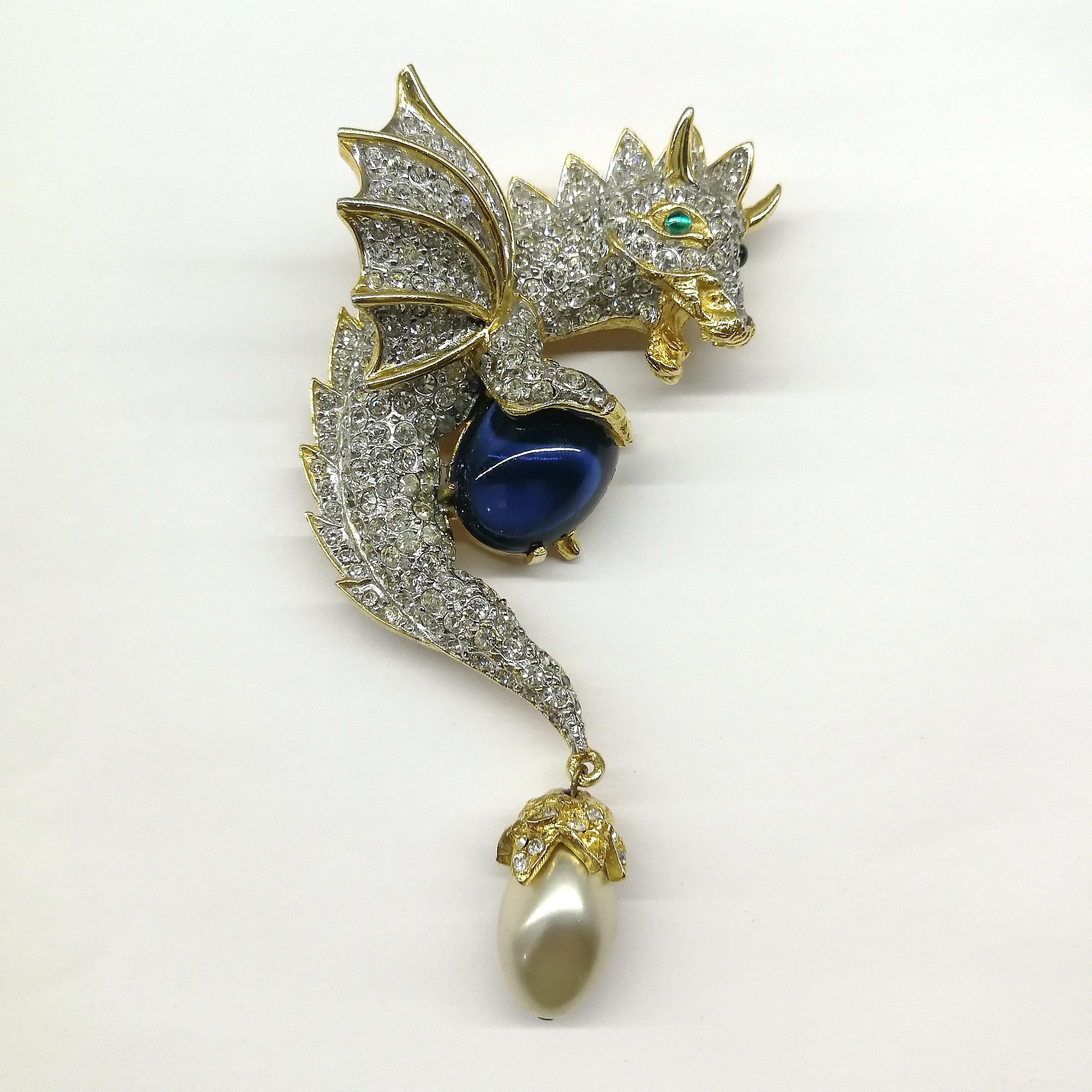 A magical and enchanting brooch from Ken Lane's heyday in the 1960's, a greedy dragon holding fast to his jewel, with a cheeky grin! With handset clear pastes, a large sapphire cabuchon 'jewel', a baroque pearl suspended from the tip of his tail,