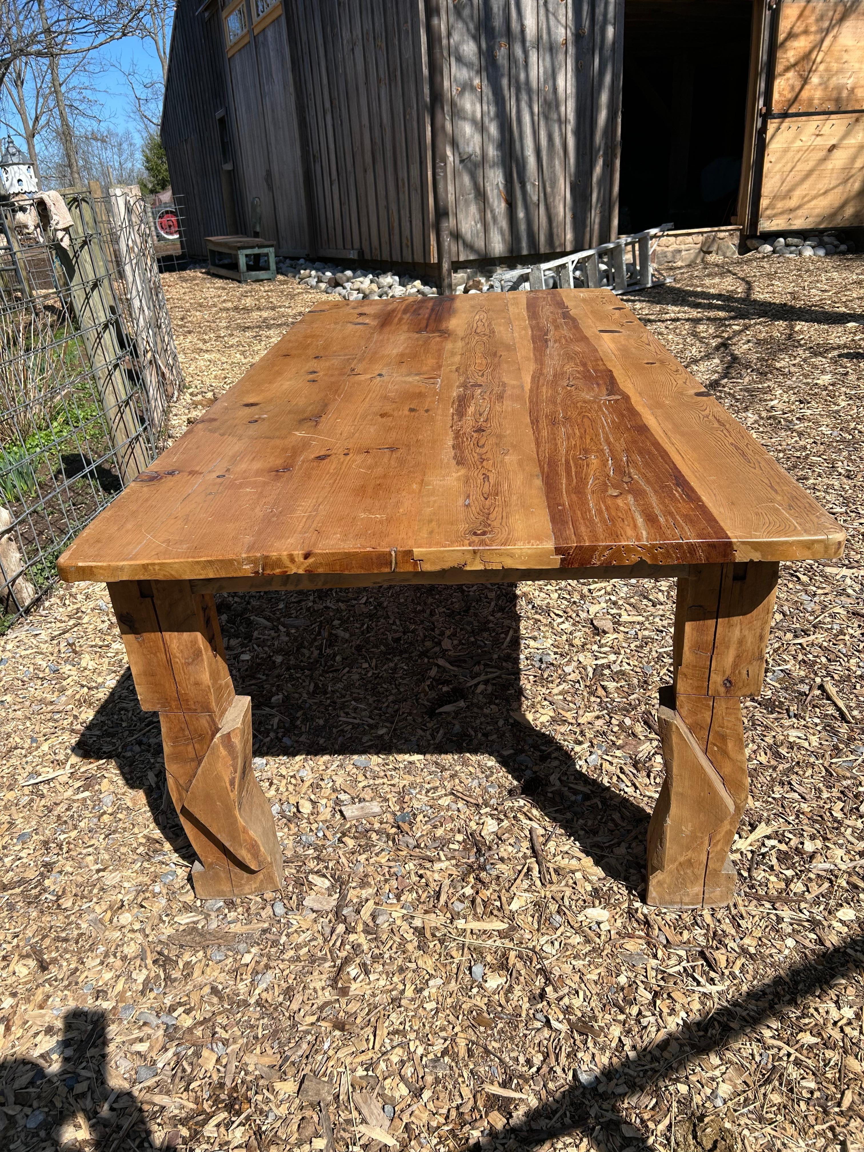 Impressionnante table de ferme rustique en pin lourd fabriquée à la main avec des pieds géométriques sculptés et de belle couleur miel naturelle avec de beaux grains et nœuds sur le dessus.  Tablier 
