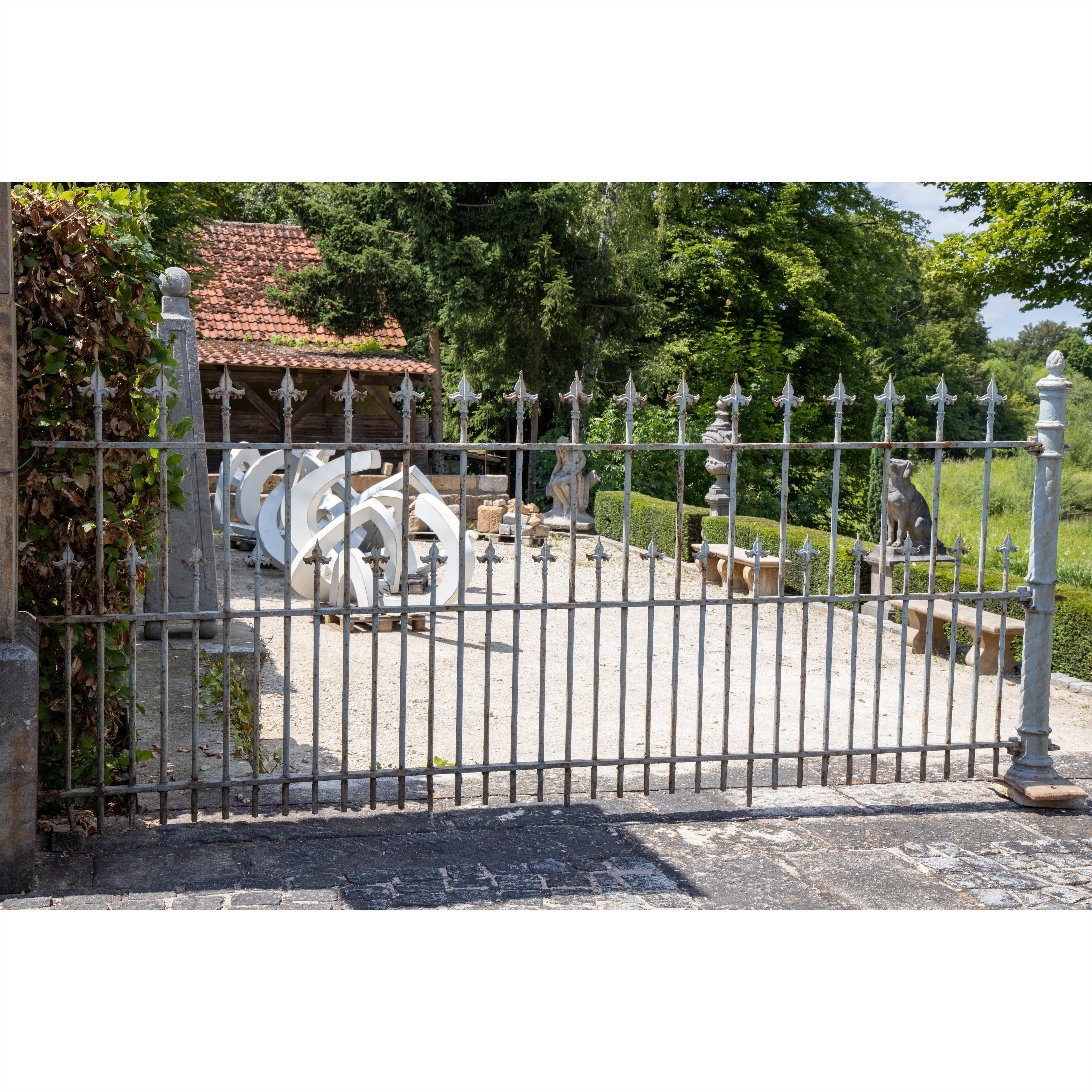 Very large, centrally raised courtyard gate with fleur-de-lis tops and ring elements, patinated in light grey-blue. Fence sections (Among others: 120 x 282 x 32 cm) and an additional door are available.