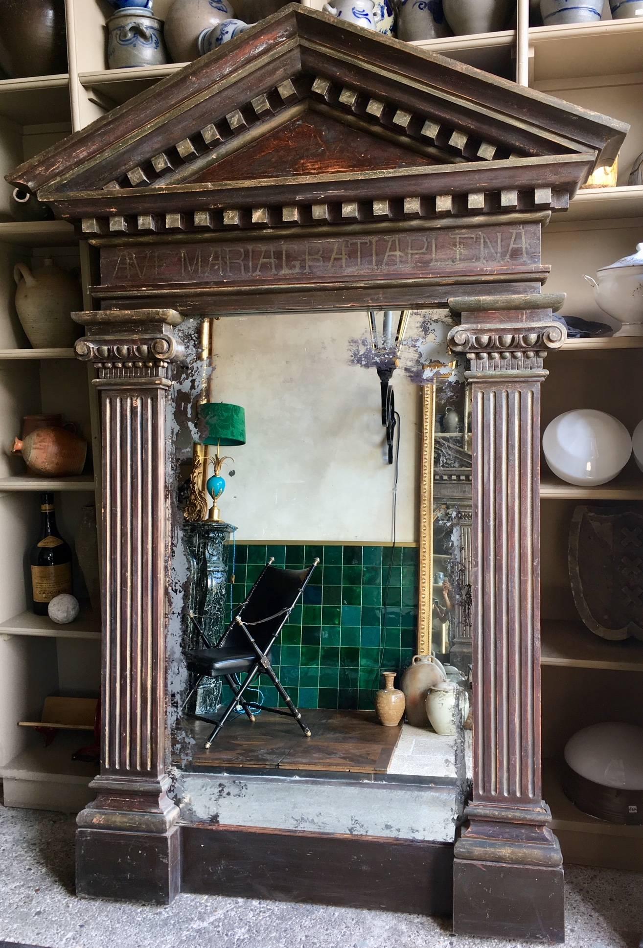 Large mirror in antique decorative frame. The frame is decorated with architectural elements such as a tympanum and Ionic pilasters. Under the tympanum the text 