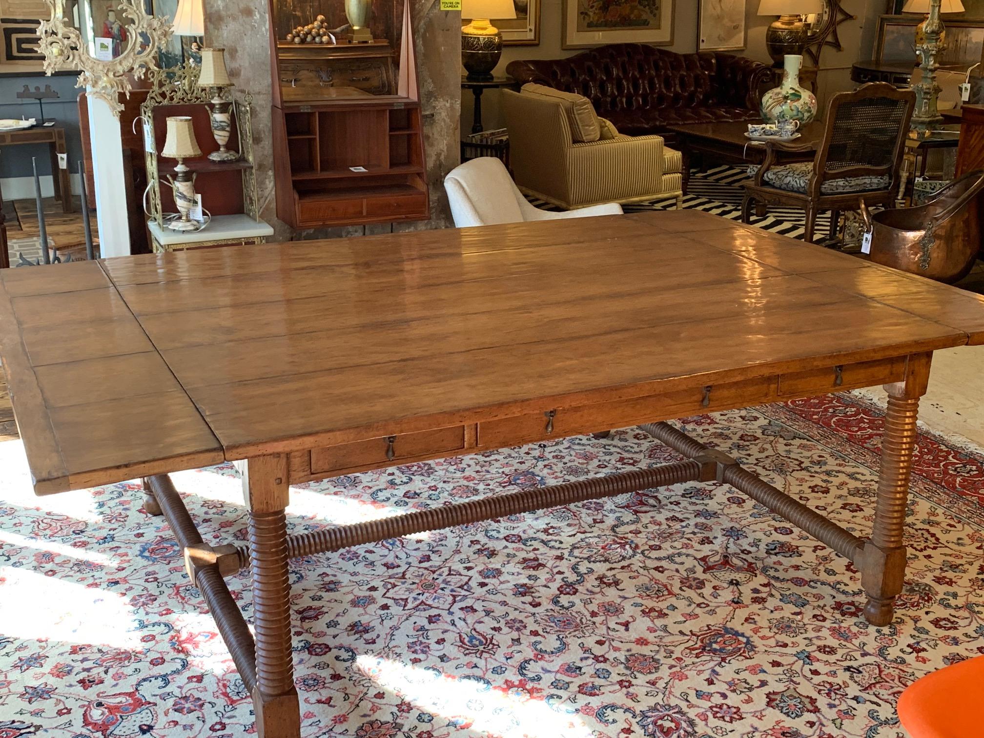 Custom center or dining table having planked walnut top, drop-leaf ends, carved screw legs, H-stretcher on ball feet, and three drawers on the frieze of both sides with antique brass Dutch-style drop pulls. Table is comparable to Rose Tarlow Melrose