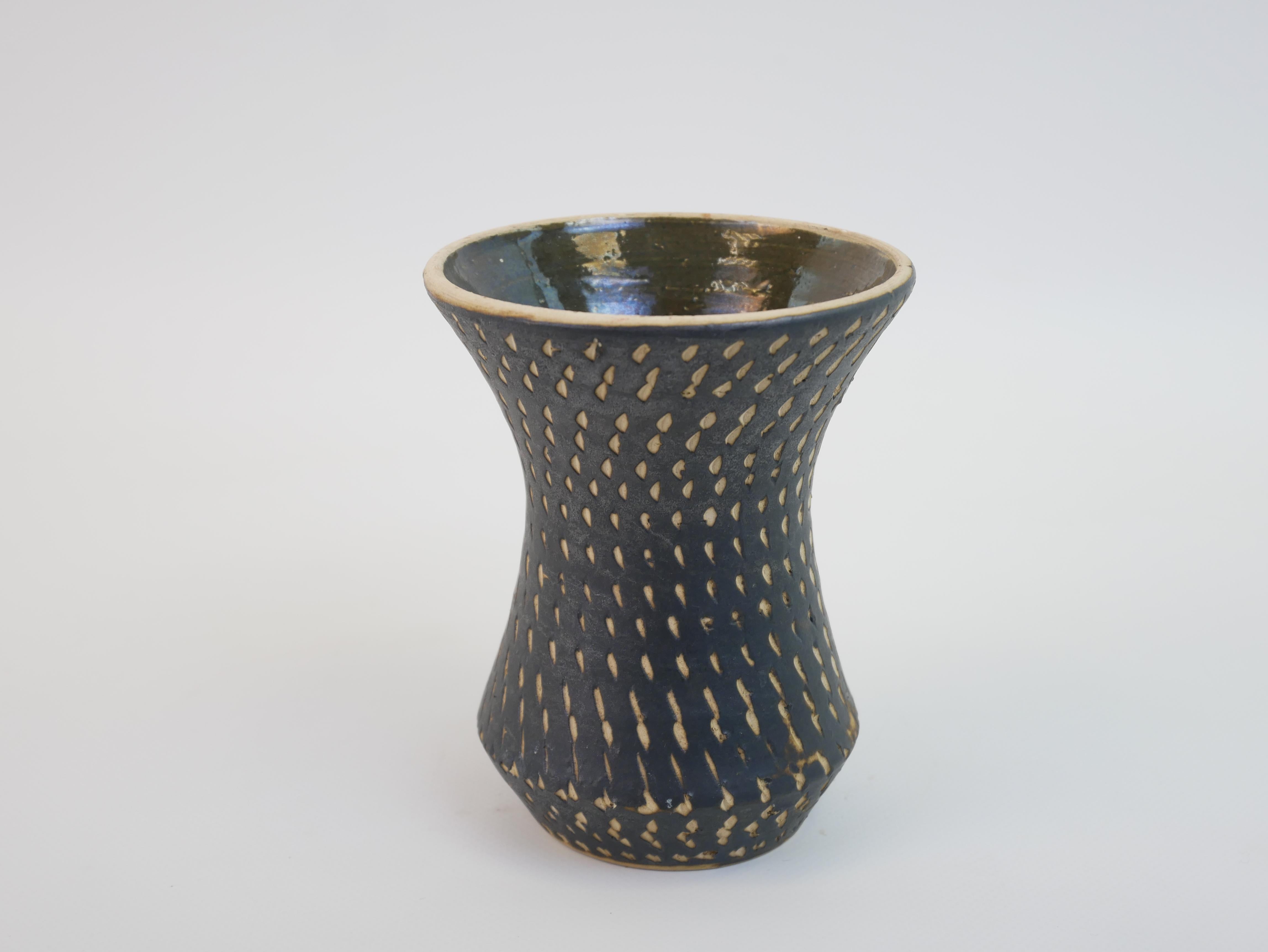 Ce vase en céramique, dans le style de Jean Besnard, présente une élégante silhouette en sablier avec une surface texturée et incisée à la main. La glaçure noire mate et profonde est ponctuée d'un motif rythmique de petites indentations allongées