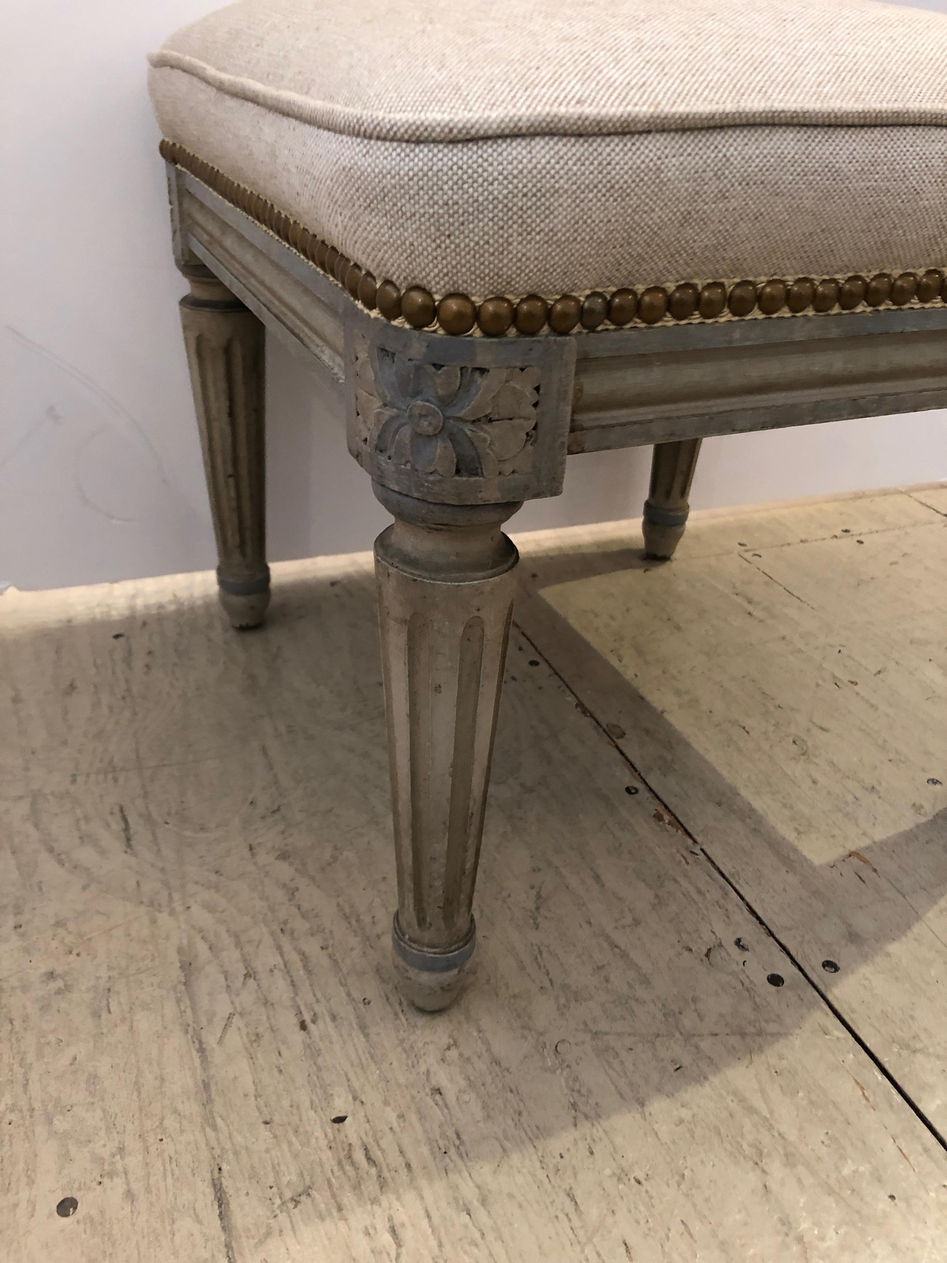 A very pretty French carved wood ottoman having original soft taupey grey paint, and new neutral Belgian linen upholstery finished with brass nailheads. Legs are fluted and tapered.