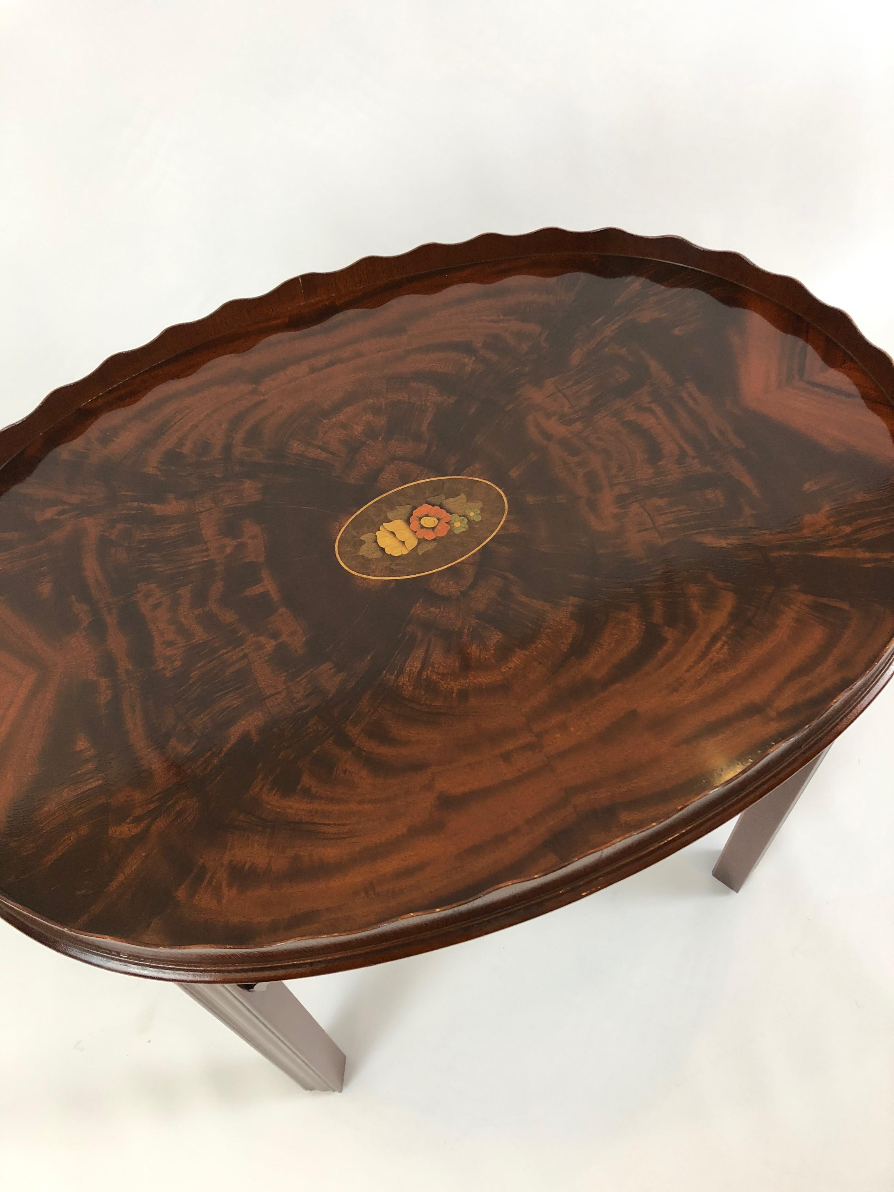 Exquisite oval cocktail table having beautifully bookmatched lively flame mahogany top with pretty inlaid floral central medallion, elegant scalloped wood edge, and refined carved wood legs.