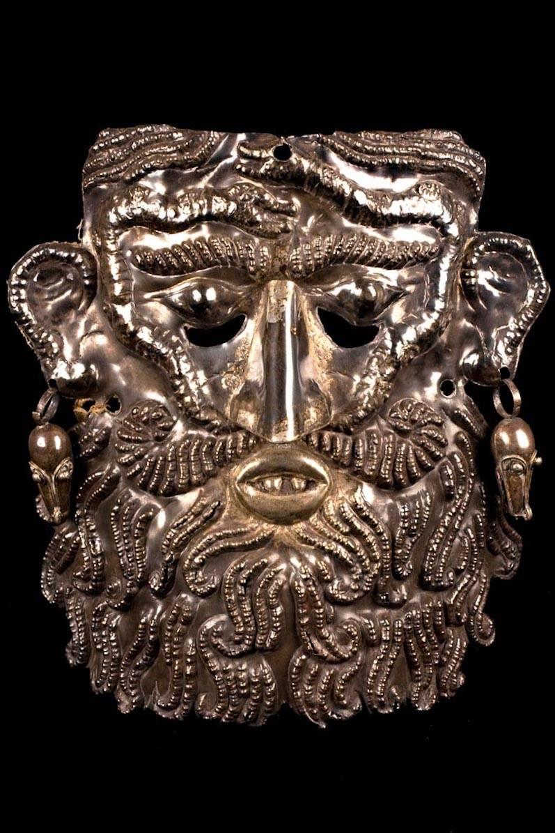 A highly decorated, heavy sheet silver mask with high relief decoration, depicting a man’s face with a long and curly beard. Two snakes decorate his forehead. The mask has pierced ears with two hollow-ware zoomorphic head earrings. Chin rest on the