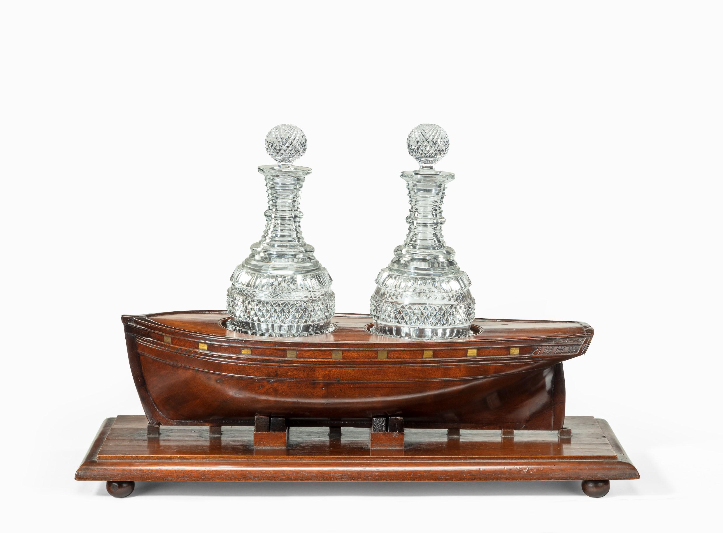 The stand for two decanters is modelled in the form of a Royal Naval ship with the original gilt gun ports. It has two circular apertures cut into the deck for a pair of cut-glass decanters and is set on dockyard building blocks. The mahogany plinth