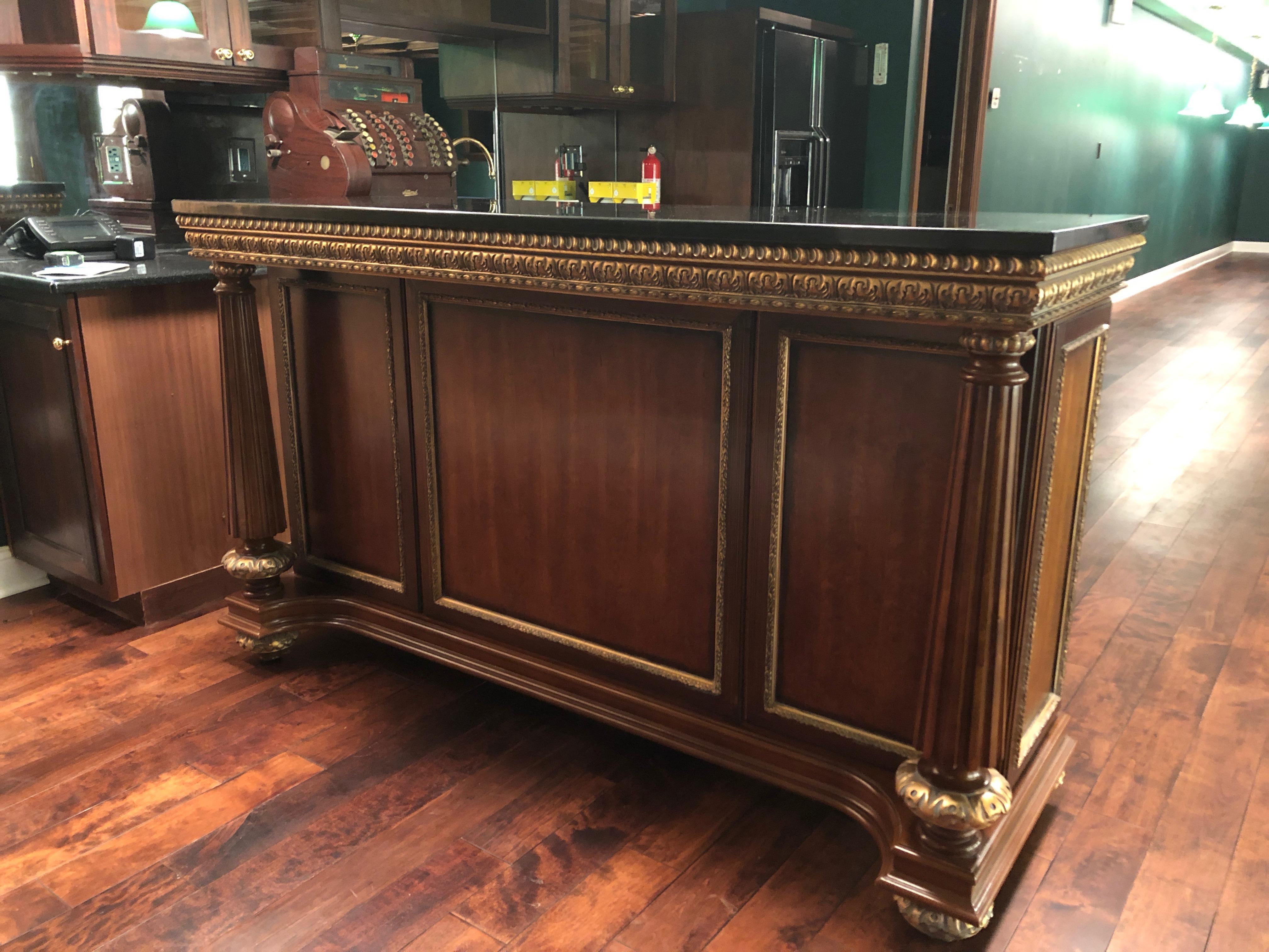A sumptuous custom dry bar cabinet having rich cherry cabinetry, fluted columns on the front and ornate gilded carved wood decorations. The top is a handsome black and white grained granite. The back of the bar has a wine rack, 3 drawers, a pull out