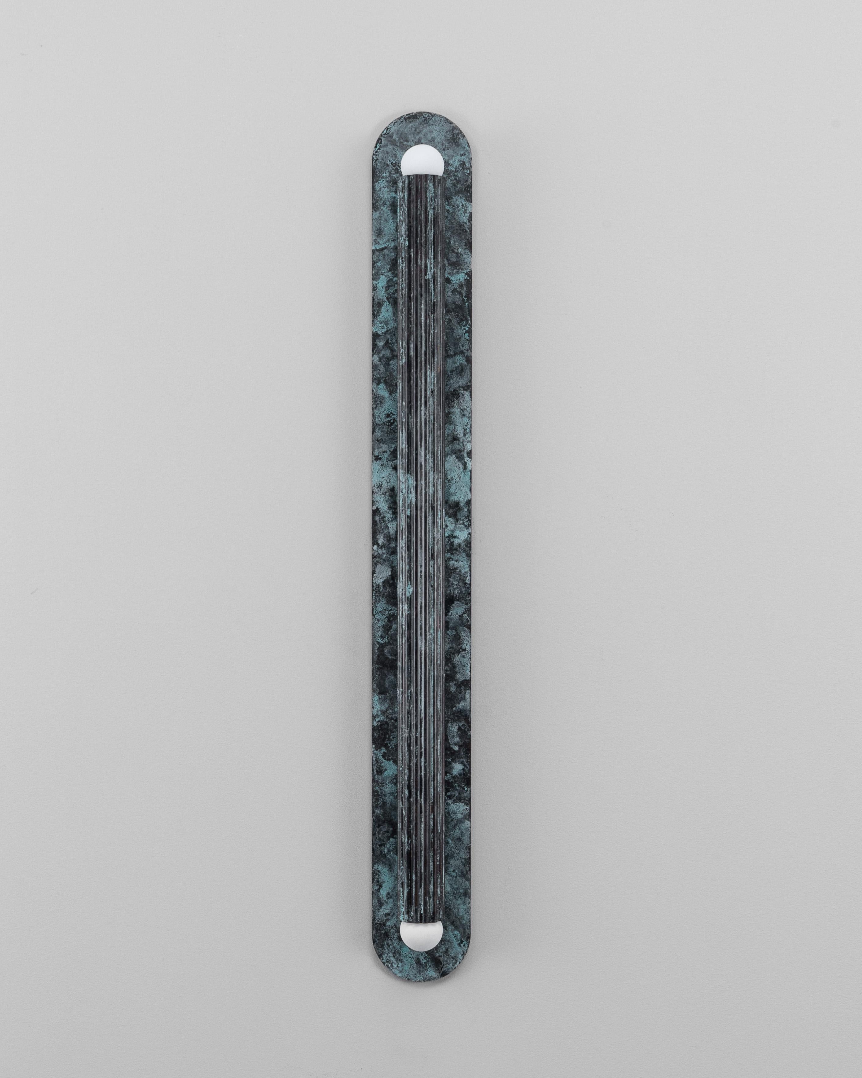 Un singolo cilindro scanalato poggia su un piatto ovale, la cui geometria è distillata in forma pura. Interamente realizzato in ottone massiccio, cattura la luce quando si increspa sulle linee verticali, trasmettendo calore, profondità e tranquilla