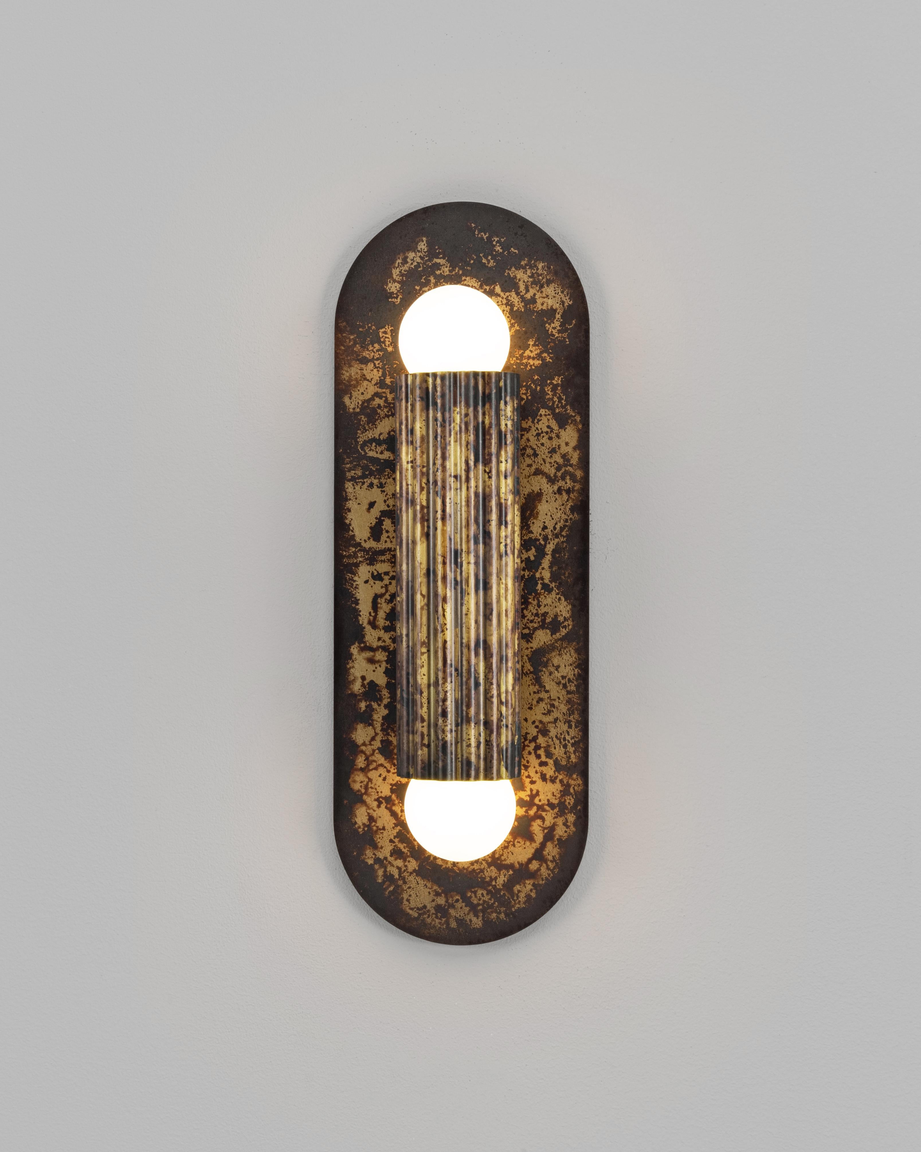 Un singolo cilindro scanalato poggia su un piatto ovale, la cui geometria è distillata in forma pura. Interamente realizzato in ottone massiccio, cattura la luce quando si increspa sulle linee verticali, trasmettendo calore, profondità e tranquilla