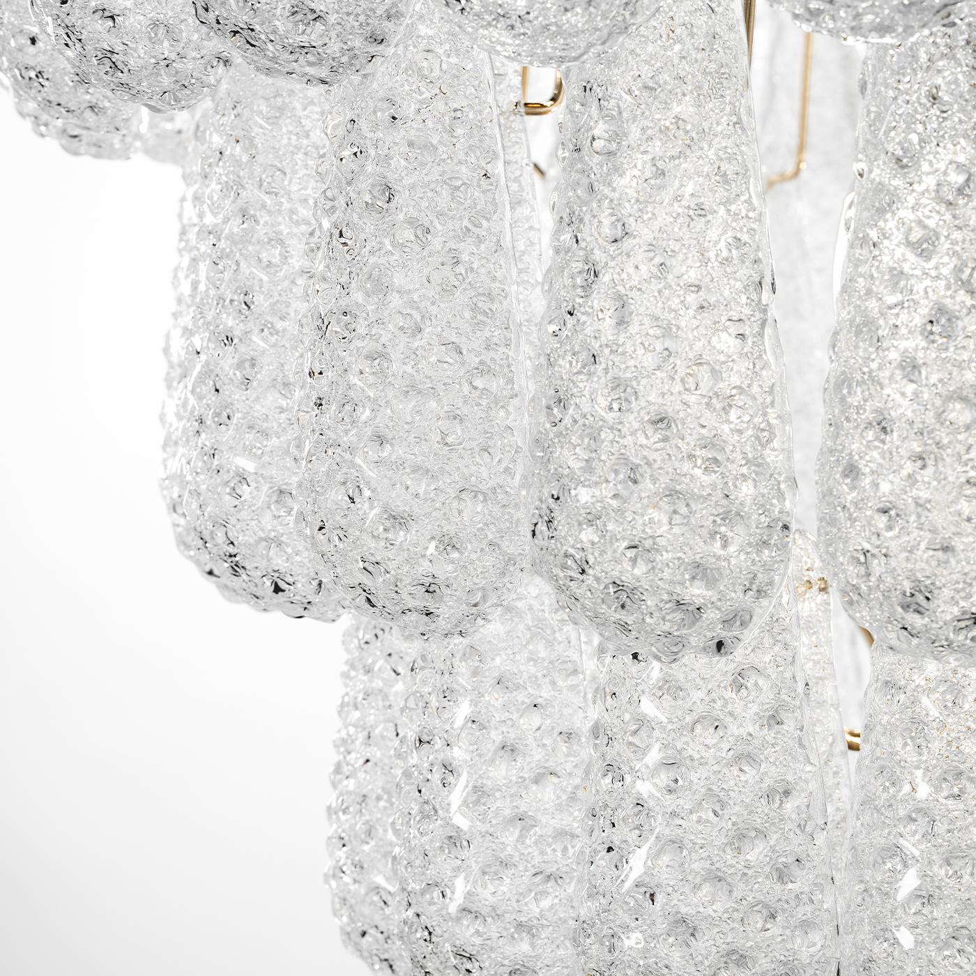 Le lustre Vetra Lux se caractérise par une armature en laiton poli supportant de multiples éléments en verre en forme de goutte dont la surface texturée diffuse la lumière pour un effet dynamique et rayonnant. Ses dimensions sont de 80 cm de