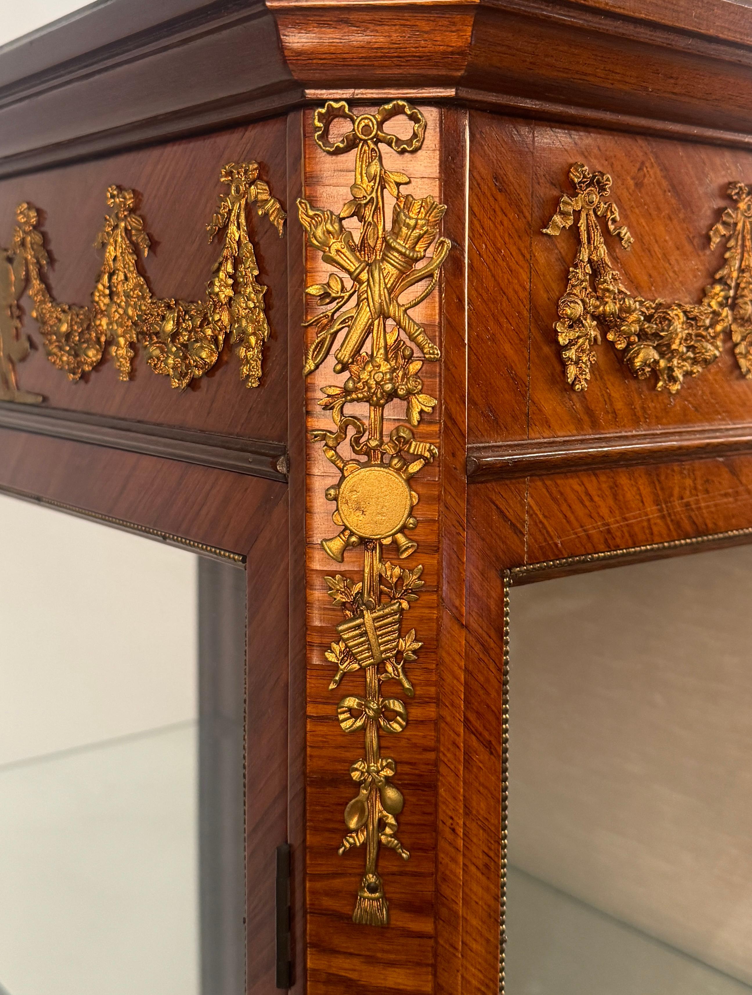 adornado con frisos de bronce dorado, frente y laterales de cristal, banda superior decorada con una danza de putti entre guirnaldas de flores repetidas en los laterales un friso de trofeos musicales en las esquinas, patas arqueadas terminadas en