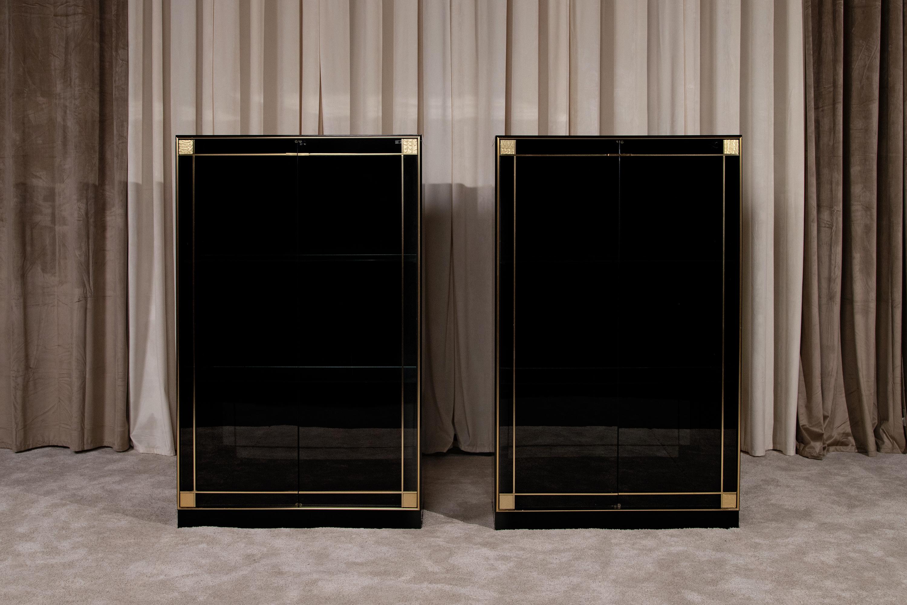 Paire de vitrines par Pierre Cardin pour Roche Bobois, production vintage des années 1980. Elles sont fabriquées en bois laqué noir brillant avec d'élégants détails en laiton dans le style Hollywood Regency. La profondeur du noir et la brillance du