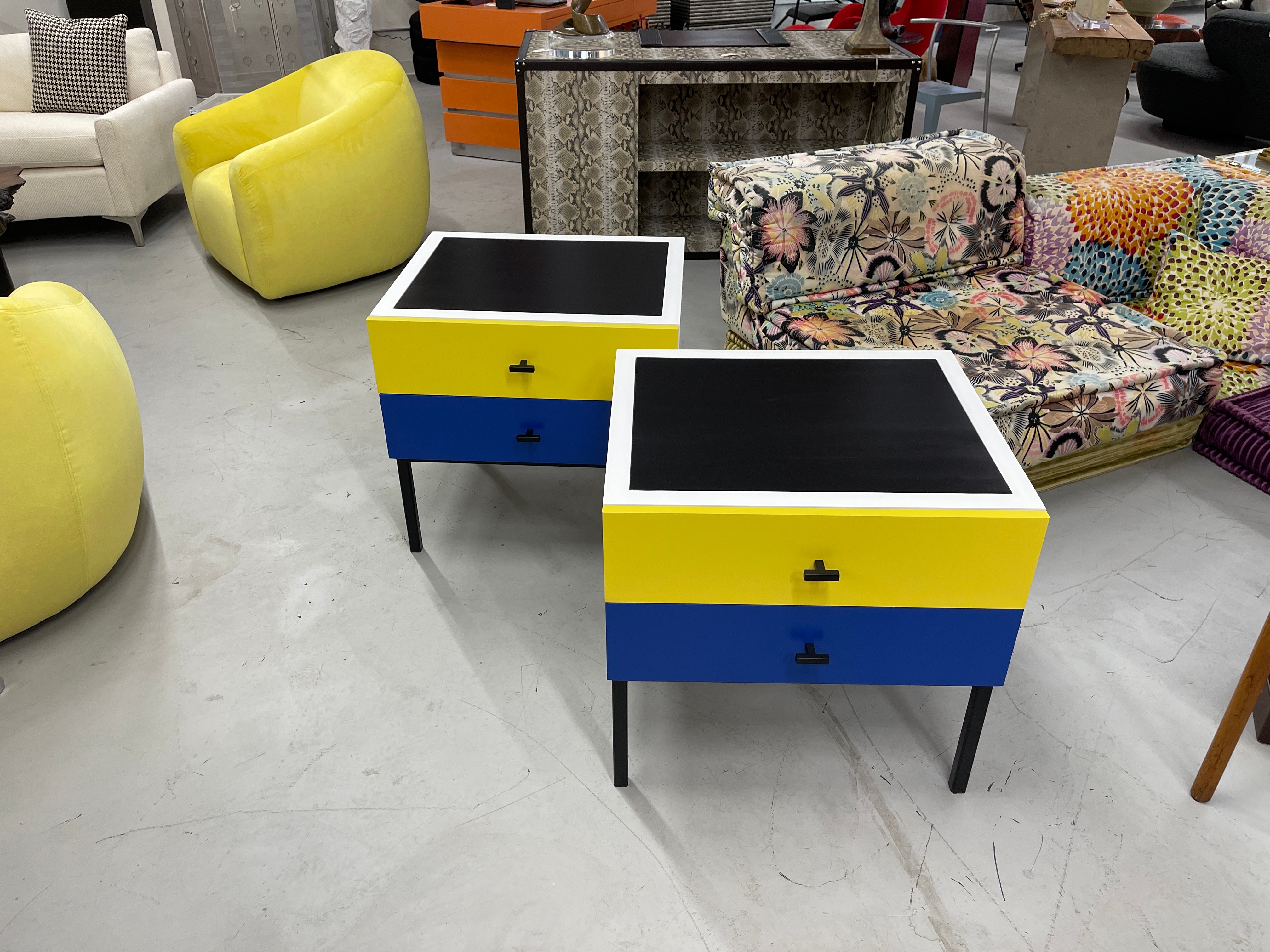 Beautiful repainted nightstands in a vibrant blue and yellow. 2 drawers each on iron legs. Inset black painted top. Nice scale and size. These nightstands are vintage pieces likely 1980's. They are 25 by 22 inches in dimension and 24 inches tall. In