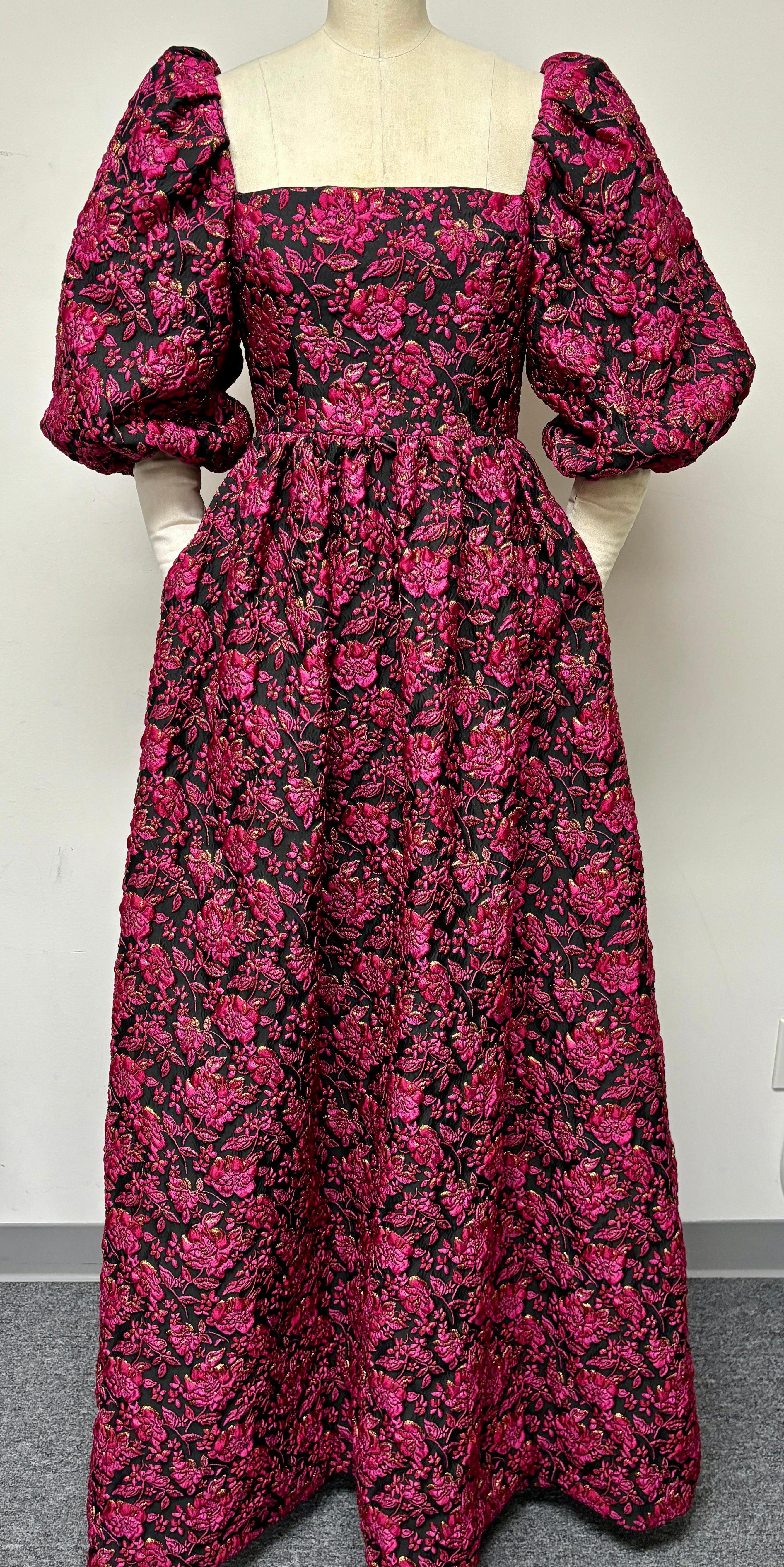 Vestido rosa y negro vibrante con detalles dorados metálicos. Un hombro con un pliegue único y una manga entera abullonada dan al vestido su exuberante plenitud y su elegancia de alta costura. El tejido con el fondo negro está salpicado de adornos