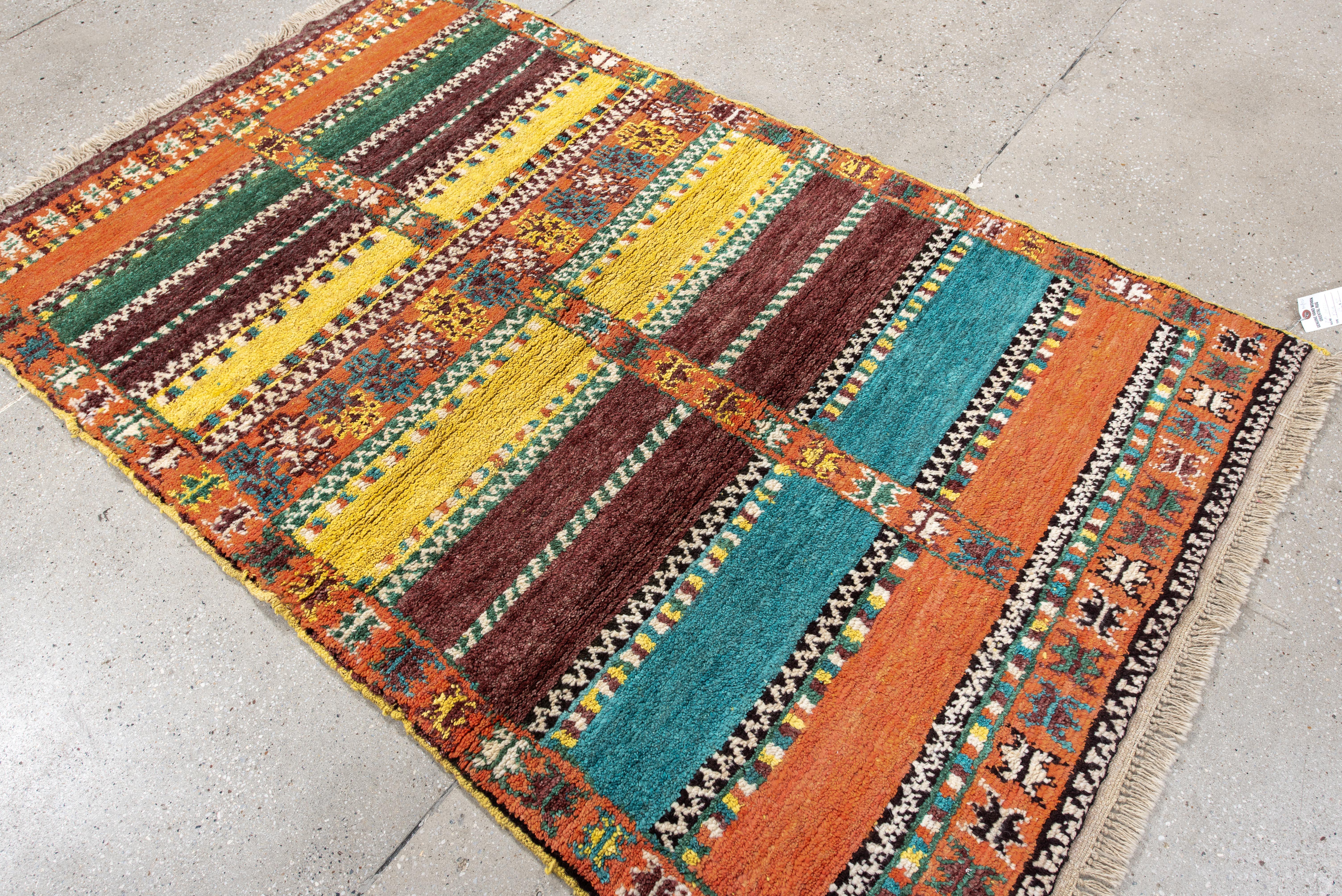 Questo vivace tappeto marocchino presenta un ricco disegno di ispirazione berbera composto da bande orizzontali alternate in ocra caldo, verde bosco, rosso rubino, giallo marigold e blu turchese. Intricati motivi tribali percorrono verticalmente la