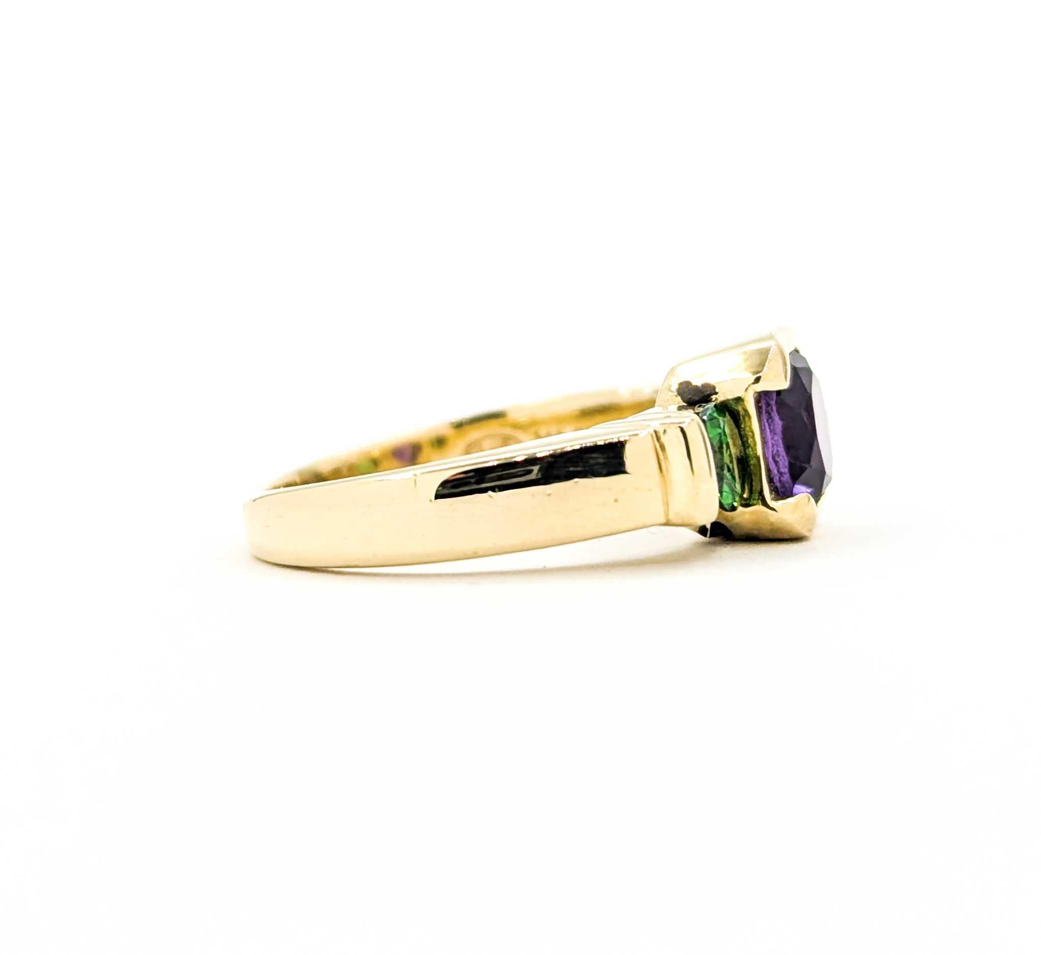 This vintage ring features a rich .75 carat purple amethyst at its center, accented on each side by bright green tsavorite garnets totaling .50 carats. The round-cut gemstones are set in secure bezels, creating a striking contrast between the royal