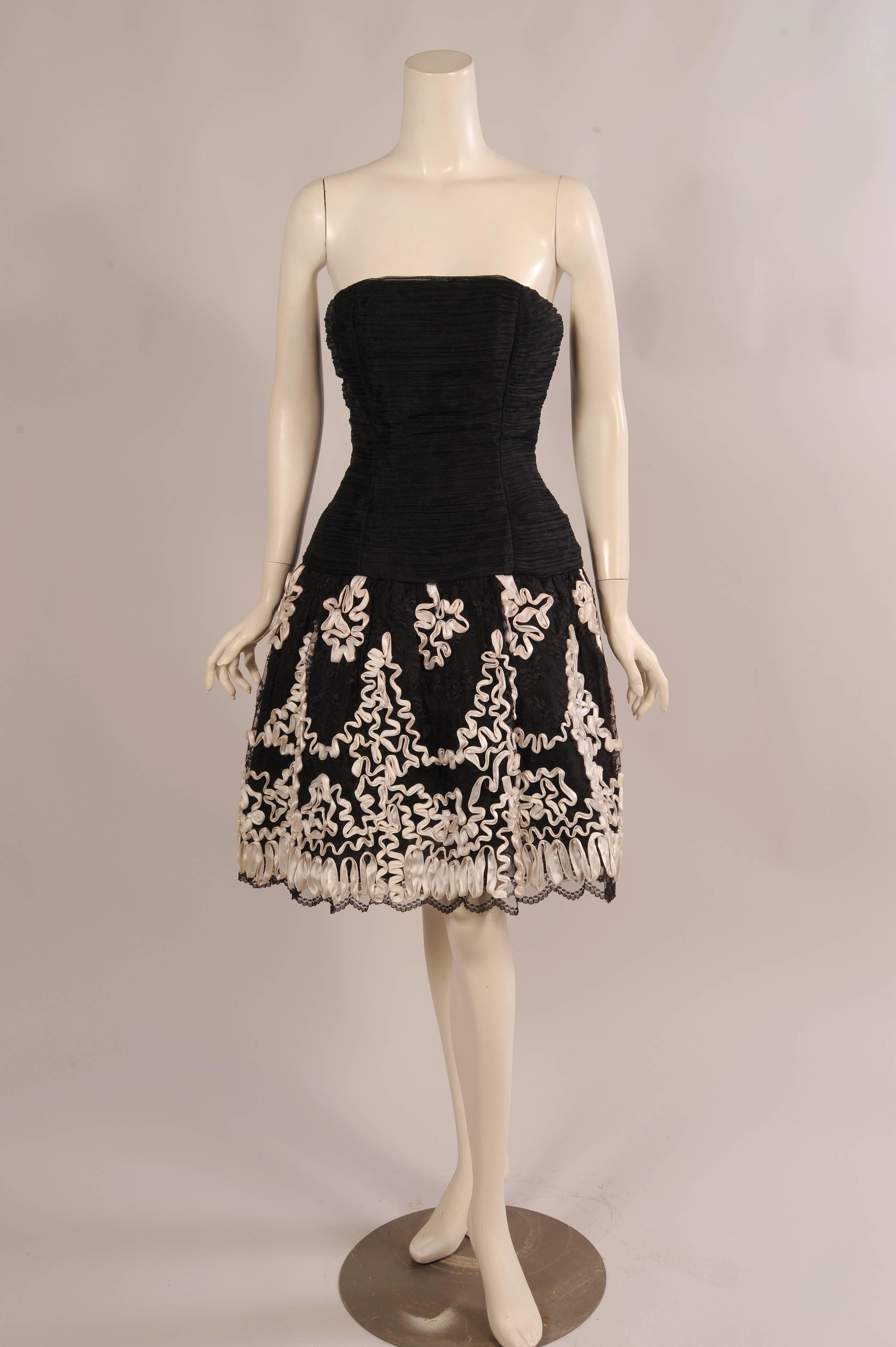 This flirty little evening dress was designed by Victor Costa for Saks Fifth Avenue. The strapless dress has a fitted organza bodice with a dropped waistline. The full skirt is black lace embellished with white satin ribbon soutache over an organza