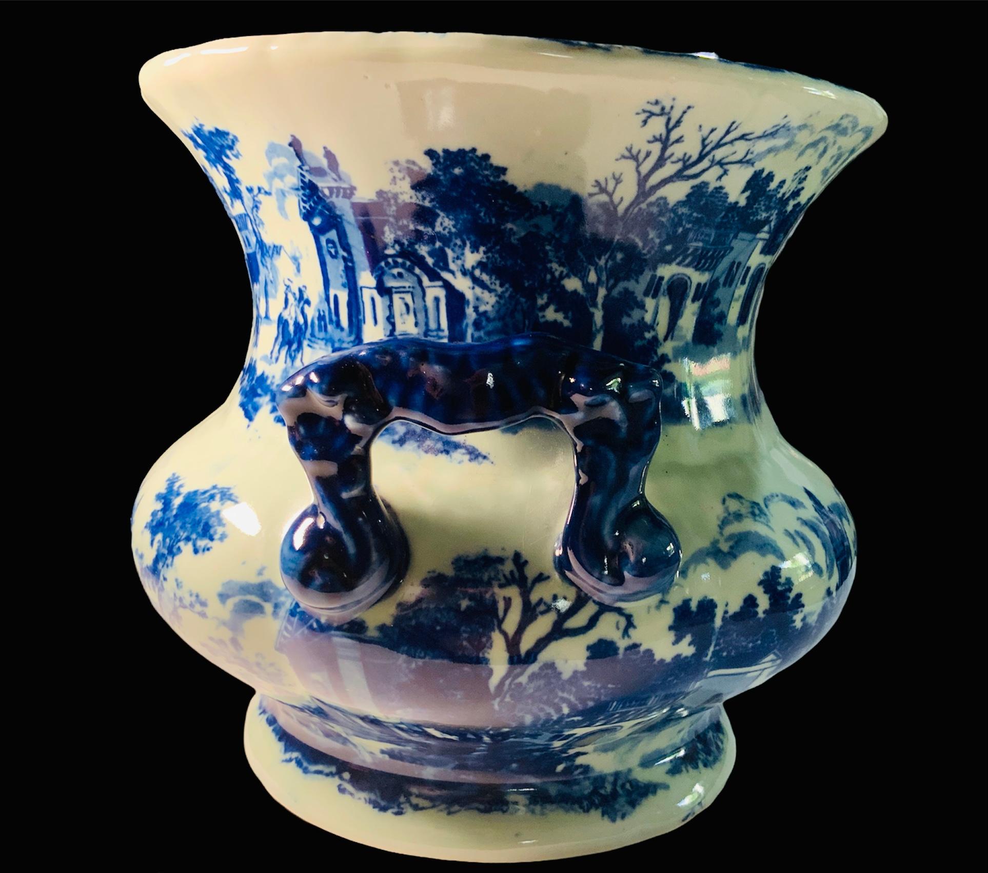 This is a Chinese reproduction of a Victoria Ware Ironstone large heavy earthenware jardiniere/planter. It is painted in white and blue with multiple flowers and foliage. It also depicts a 19th century European square scene with people walking