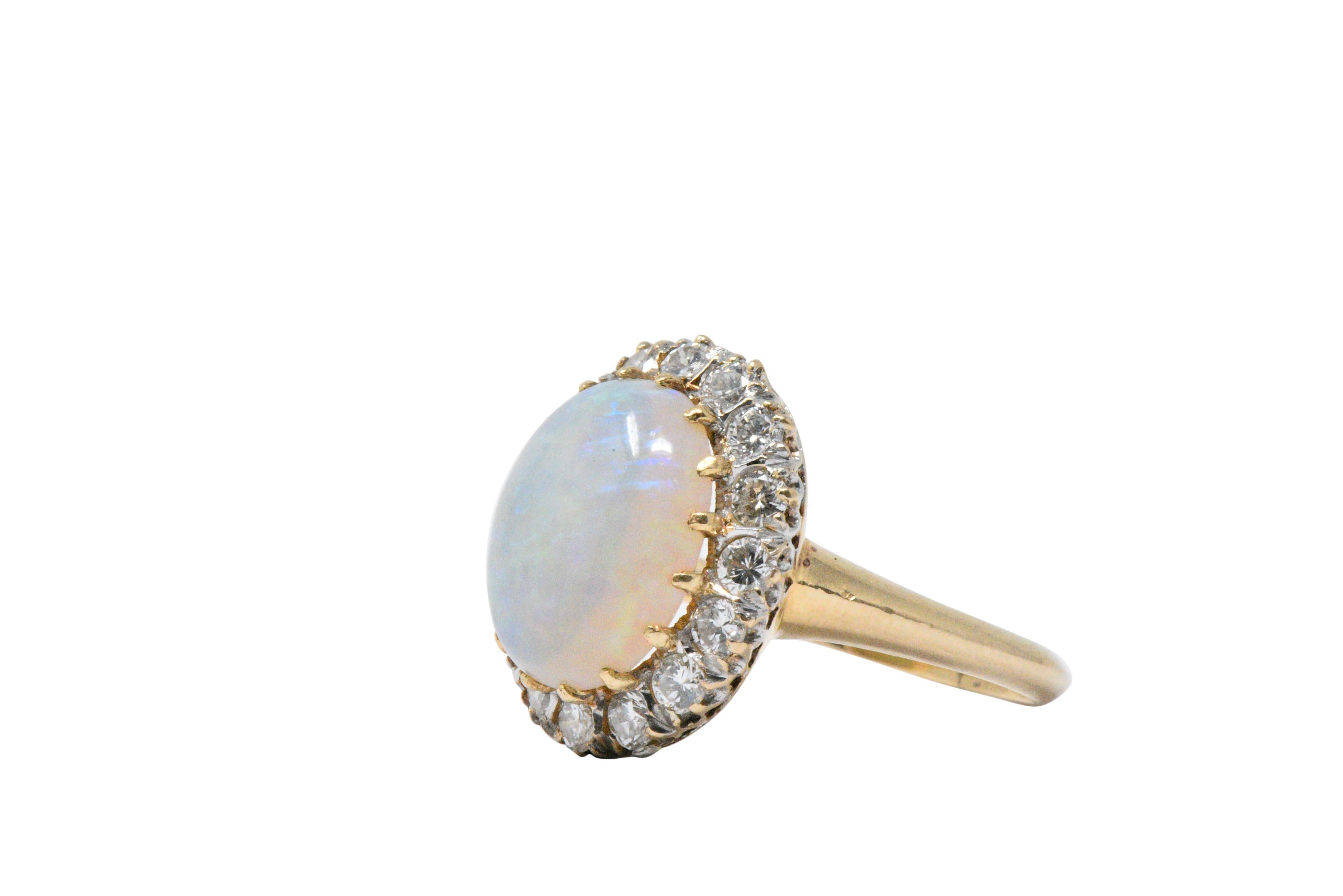 Centering an oval cabochon white opal, good play-of-color, mainly in the yellow, blue, violet and green, very good polish and luster with no signs of crazing

With a round brilliant cut diamond surround, approximately 0.65 carats total, G/H color