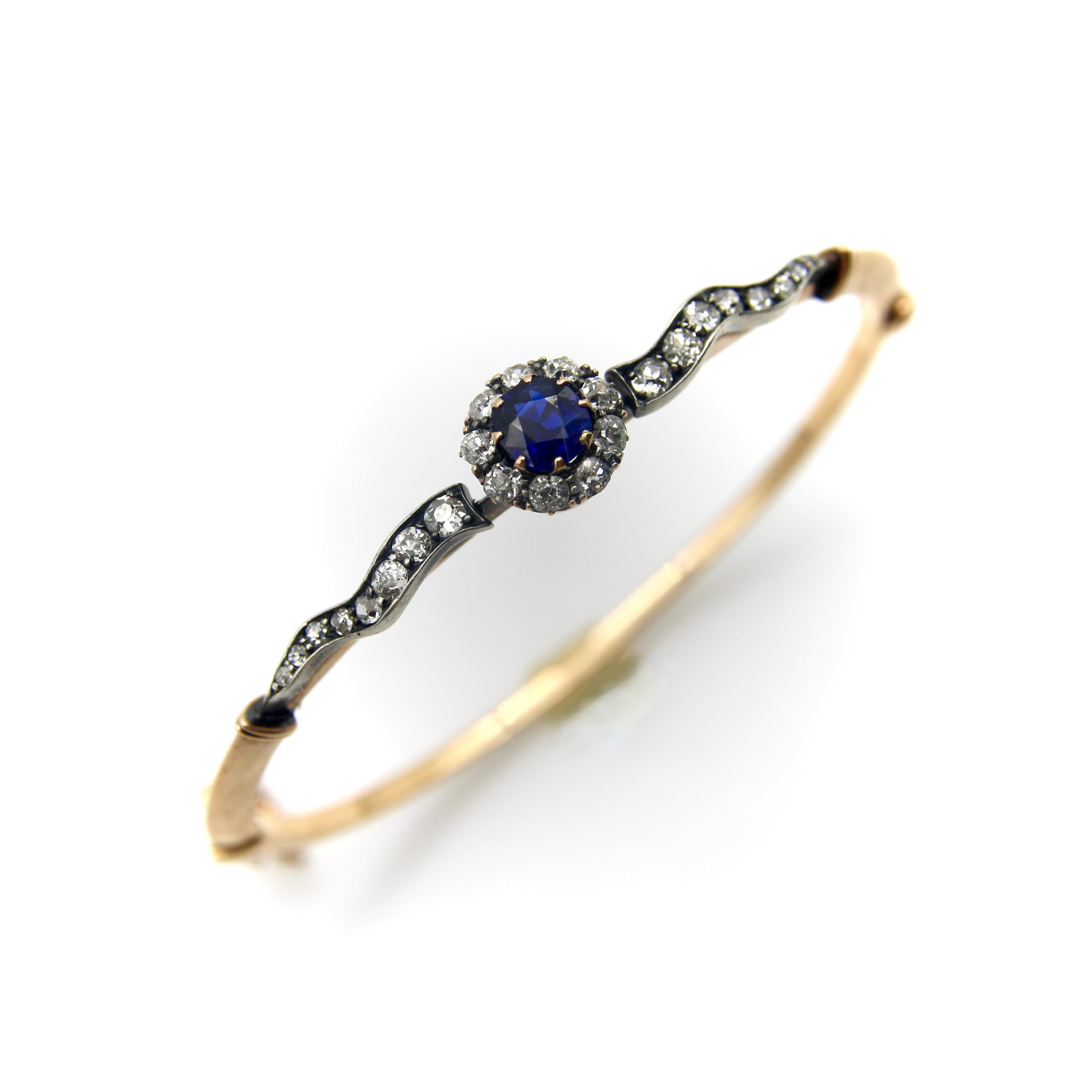 This elegant sapphire and diamond Victorian bracelet contains a saturated dark blue sapphire surrounded by Old Mine Cut diamonds. It is flanked by diamond encrusted wavy banner-like ribbons along the band. The central element is reminiscent of a