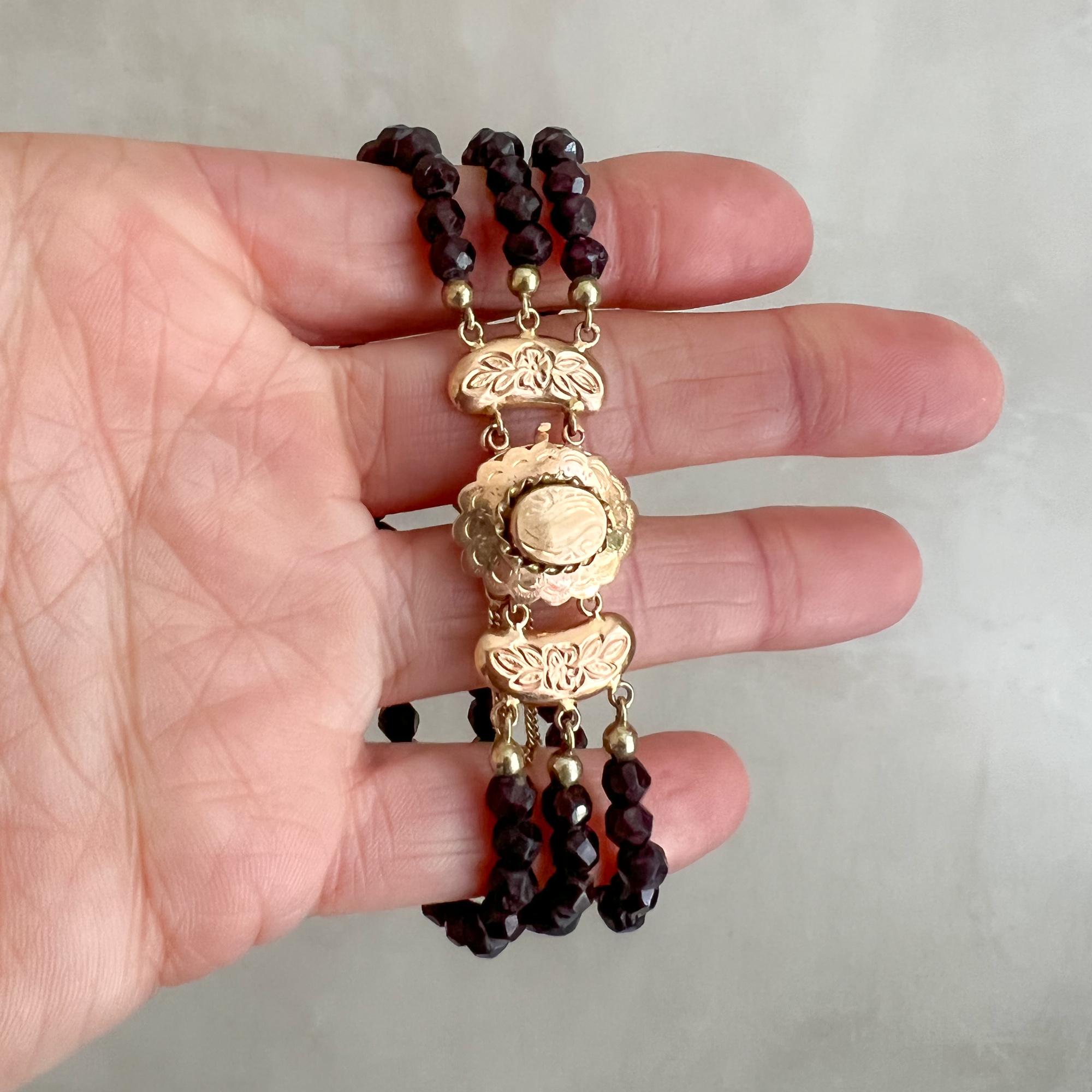 This is an antique three-strand 14 karat yellow gold garnet bracelet. The oval-shaped clasp of this bracelet is beautifully engraved with a floral design, detailed figures and a scalloped border. The raised center of the clasp is surrounded with a