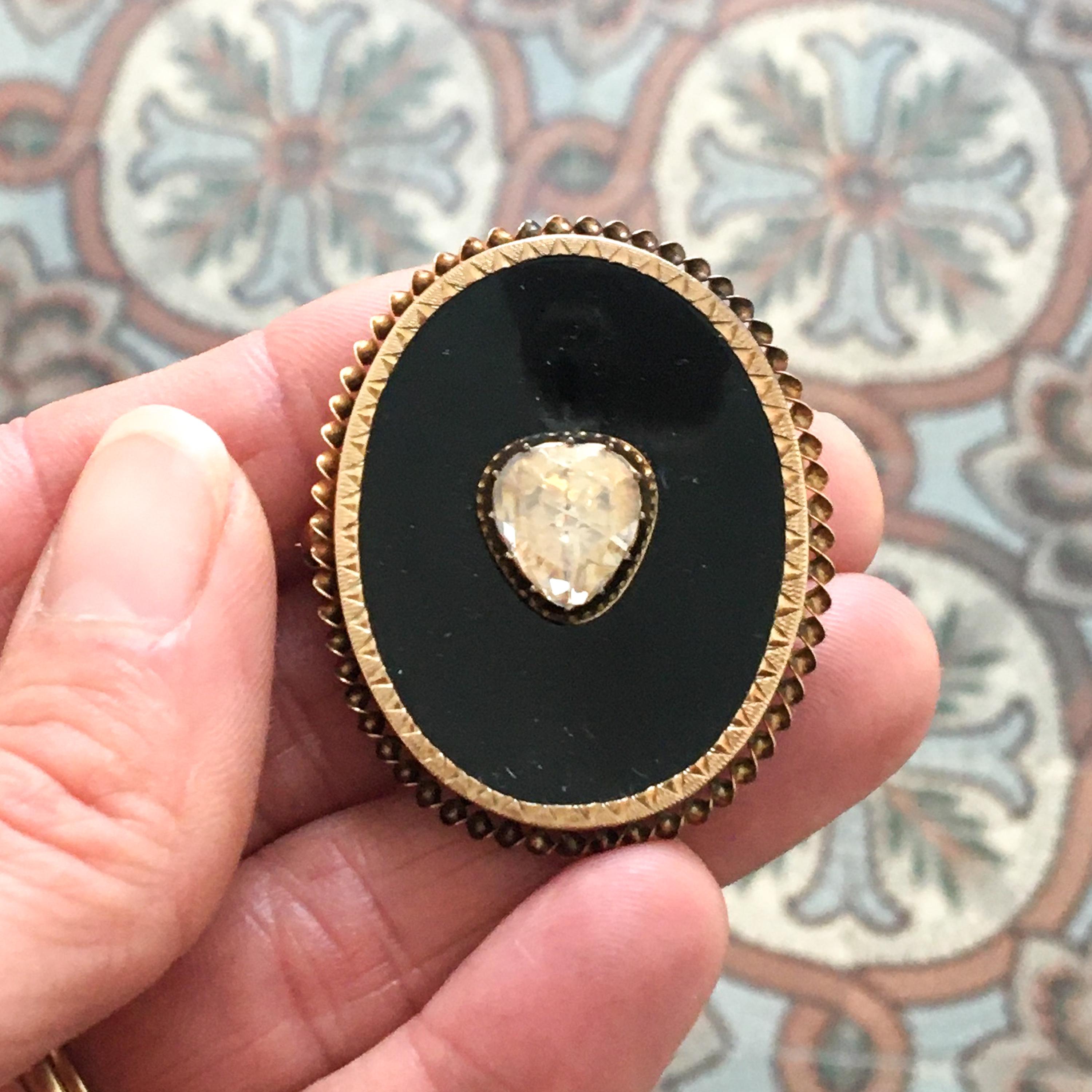 An antique Victorian rose cut diamond and onyx oval pin brooch. The onyx gradually slopes towards the diamond in the center. The heart-shaped diamond is rose cut and bezel set with small prongs on top. The 14 karat gold rim is decorated with small