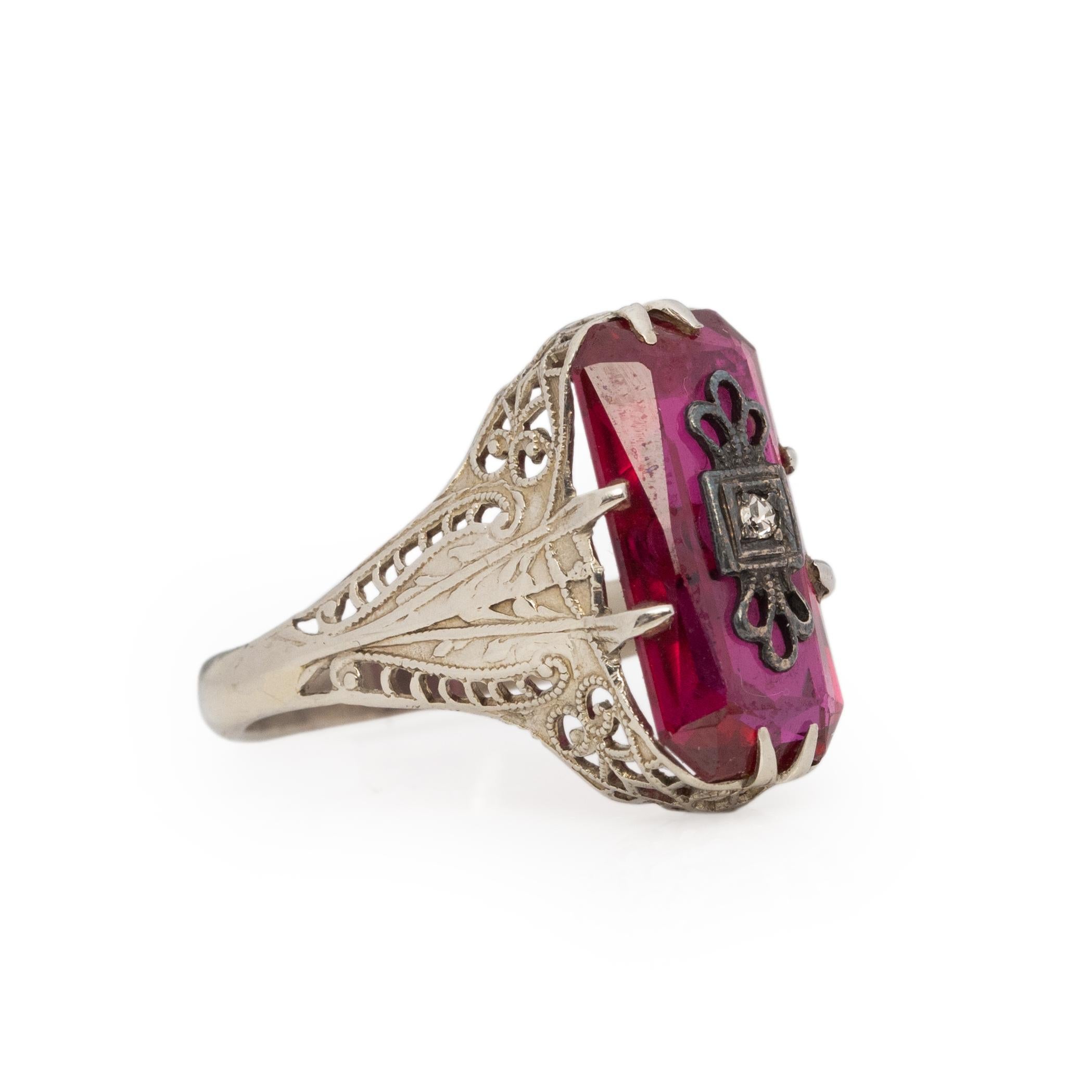 This ring is from the Victorian era, crafted in 14K white gold with intricate filigree details along the shanks and gallery. Held in by 7 prongs the rectangle shaped deep red stone center has a small diamond detail affixed to the center. Giving off