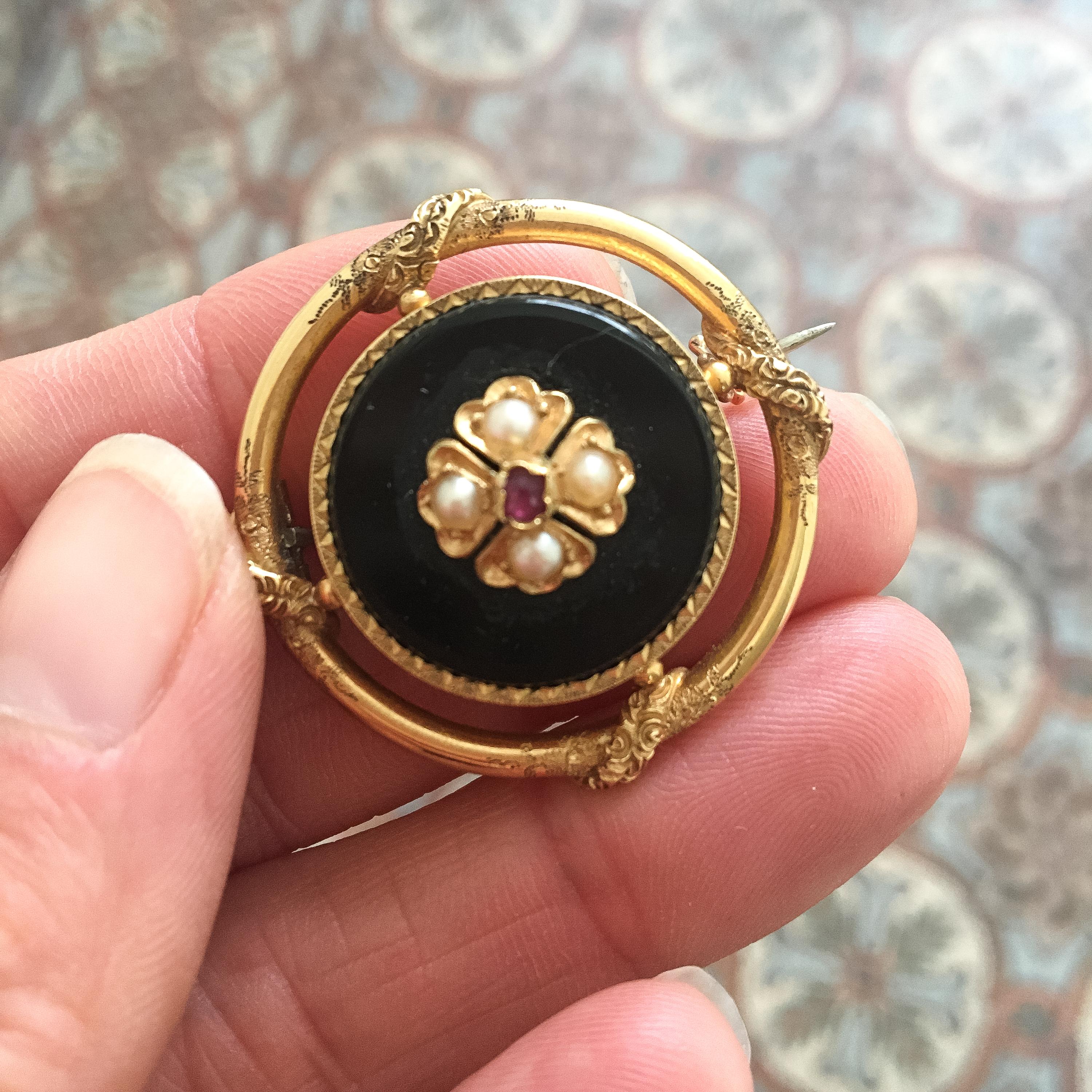 A Victorian 14 Karat gold black round onyx, seed pearl and ruby pin brooch. The round onyx stone is designed with four seed pearls and a ruby, set in a floral decor. The 14k gold border has an etched design and the gold ring around has beautiful