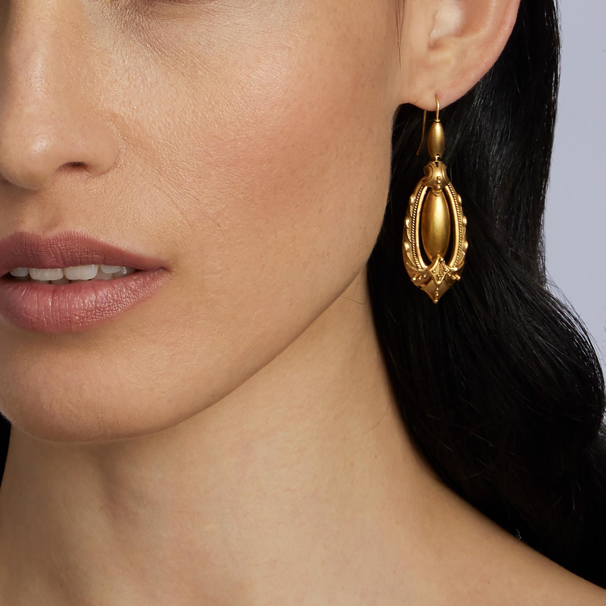 Created in the 1870s, these Victorian pendant earrings are composed of 15K gold. Each pendant earring is designed as an elongated domed oval and hoop with scroll, applied bead and wire motifs, centering a flexibly-set fully rounded oval drop.