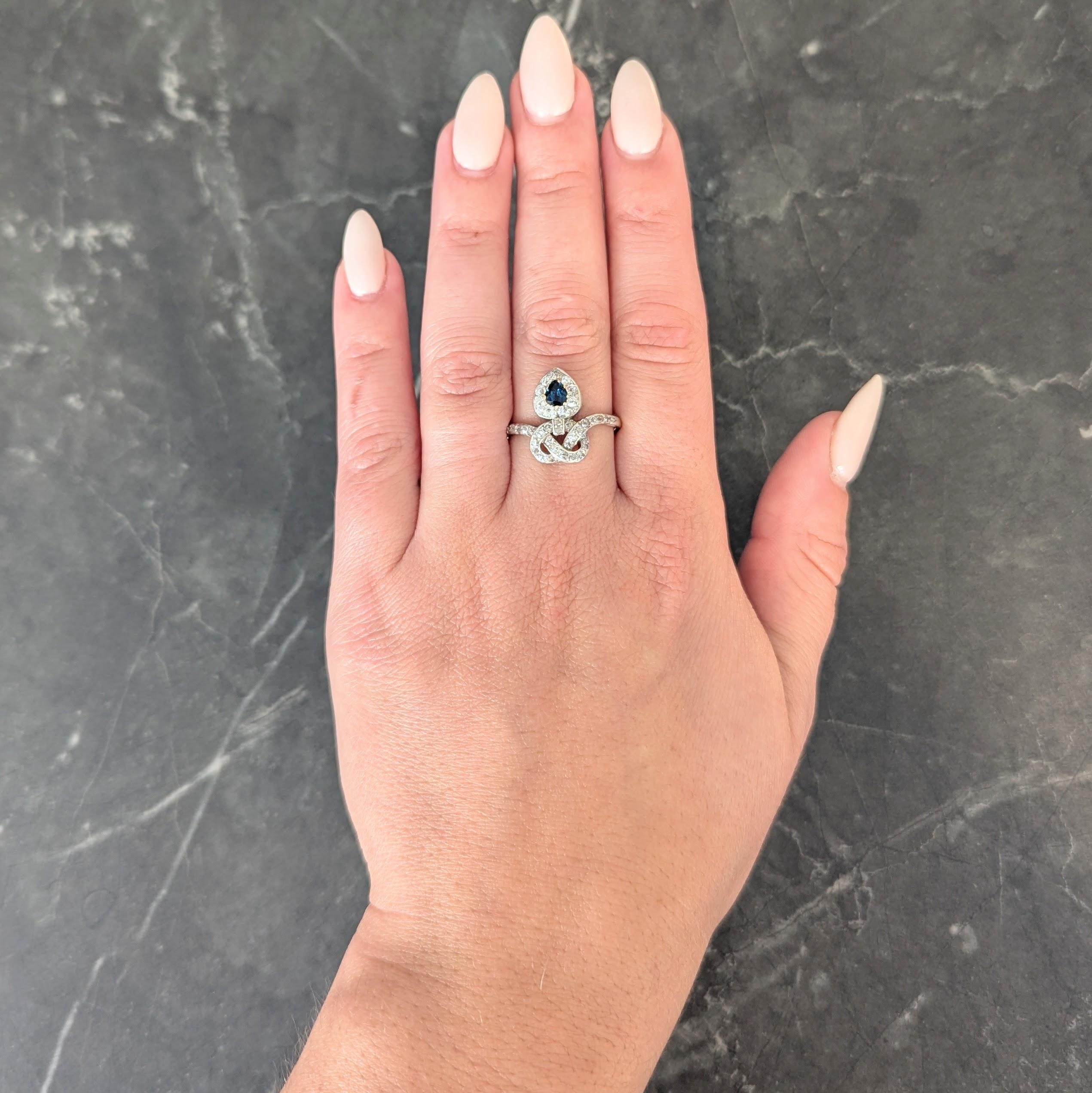 Designed as a silver-topped knot motif wrapping around a heart shaped form
Heart centers a pear cut sapphire weighing approximately 0.45 carat
Transparent medium blue in color and bead set with halo surround
Comprised of bead set old European and