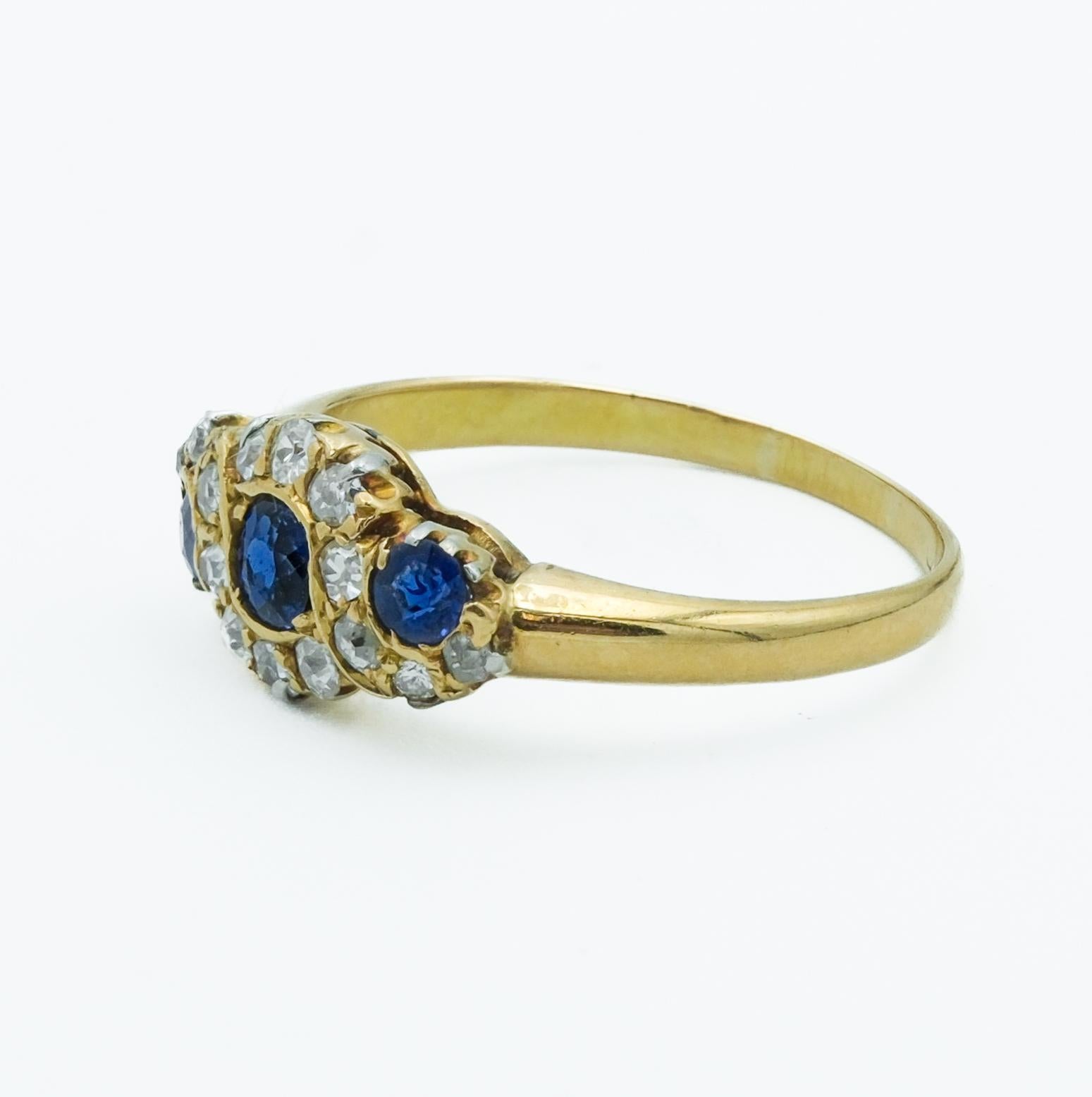 This Victorian-era ring, crafted around the 1880s, is an exquisite example of the period's intricate and opulent designs. The ring features a central royal blue sapphire flanked by two smaller sapphires, each set within a halo of diamonds. The deep