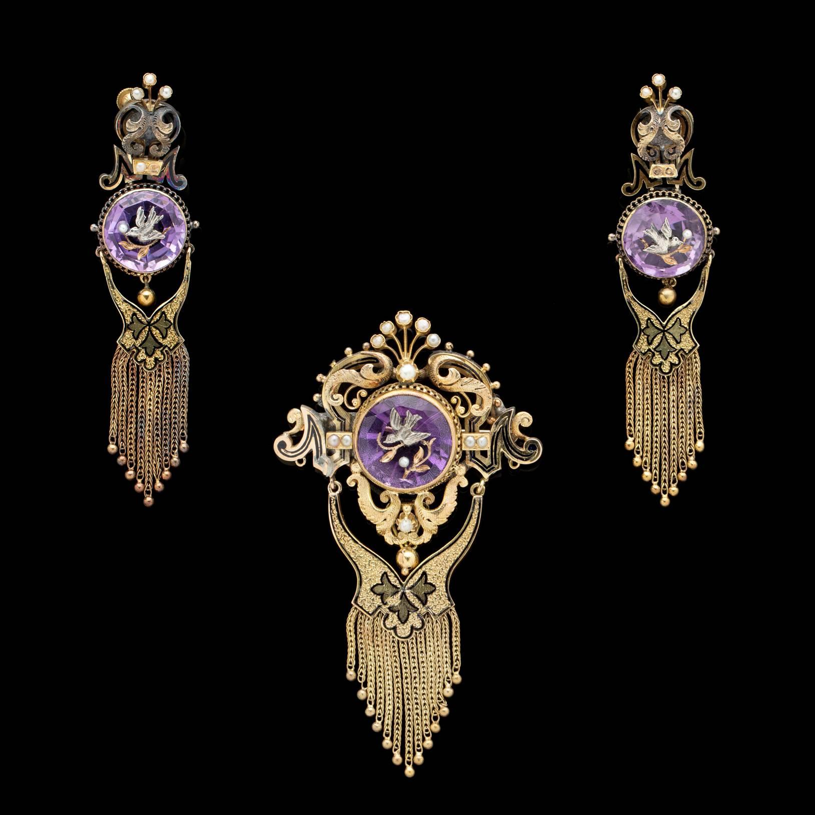 Own a piece of history. A unique 14k gold brooch and earring set circa 1870's featuring rich purple amethyst intaglios inlaid with doves and olive branches, representing hope and forgiveness. Accented with seed pearls, enamel tracery and fringe, the