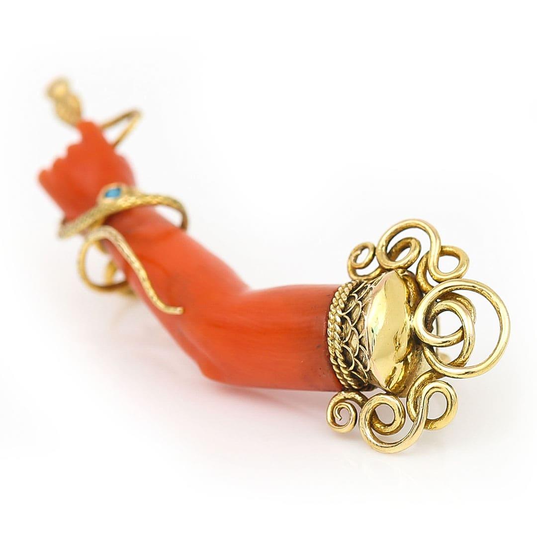 Dating from the height of the Victorian era this wondrous 18ct gold coral figa or mano figa brooch is ornately decorated in a writhing serpent, turquoise detailed bangle and ornate cursive terminal. 

The figa hand charm is a powerful symbol that
