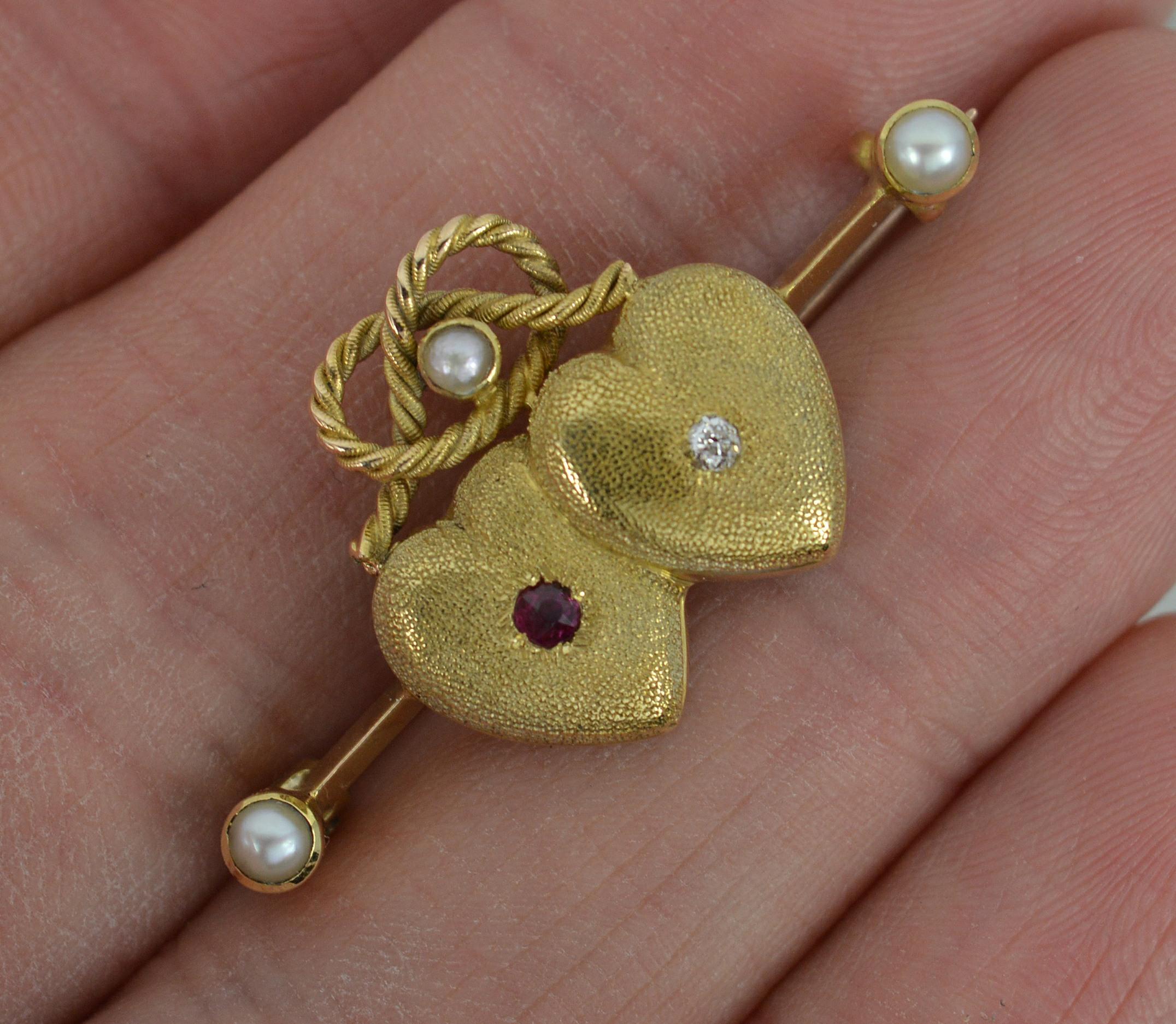 A true mid to late Victorian era brooch. c1870.
Solid 18 carat yellow gold example.
Designed with two hearts with a mottled finish. Above a knot set with pearl and a bar to each side with a further pearl. 
Set with an old cut diamond and ruby to the