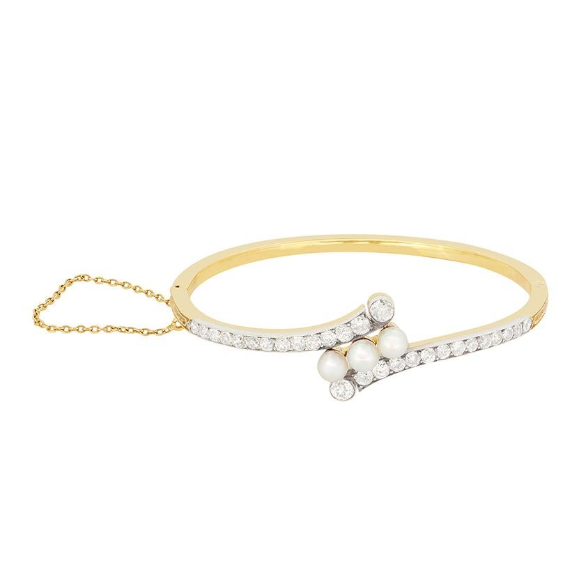 Dating to the Victorian era, this beautiful bangle features a collection of pearls and old cut diamonds. At its centre sits a line of three natural pearls. Each 5mm in diamond and set diagonally. The twisting band envelops the pearls and is adorned
