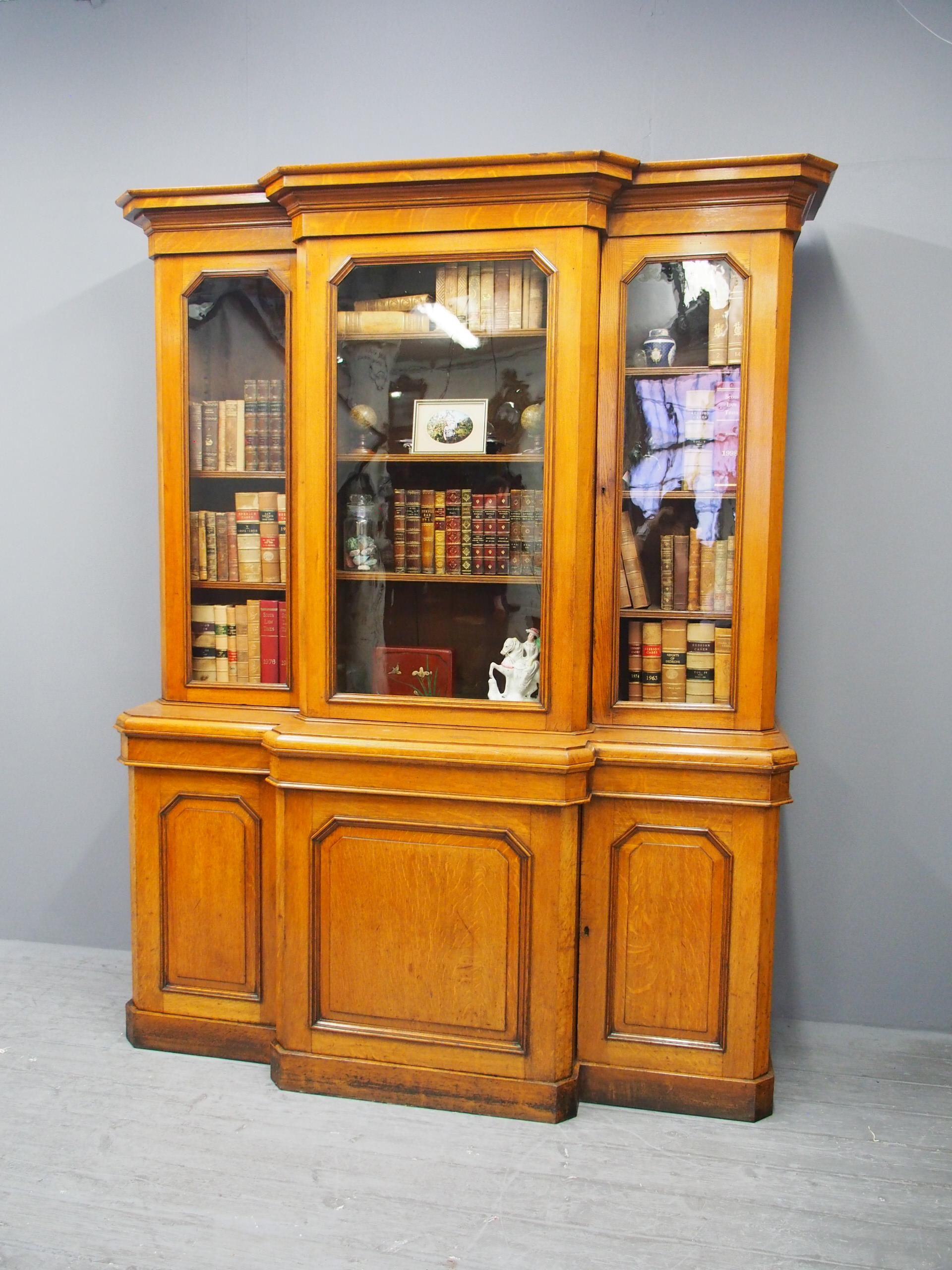 Mid-Victorian 3-door breakfront bookcase in figured quarter-cut, golden oak. The molded cornice is over a figured oak frieze and 3 glazed doors with chamfered sides, opening to interiors with oak-faced adjustable shelves. The conforming base has a