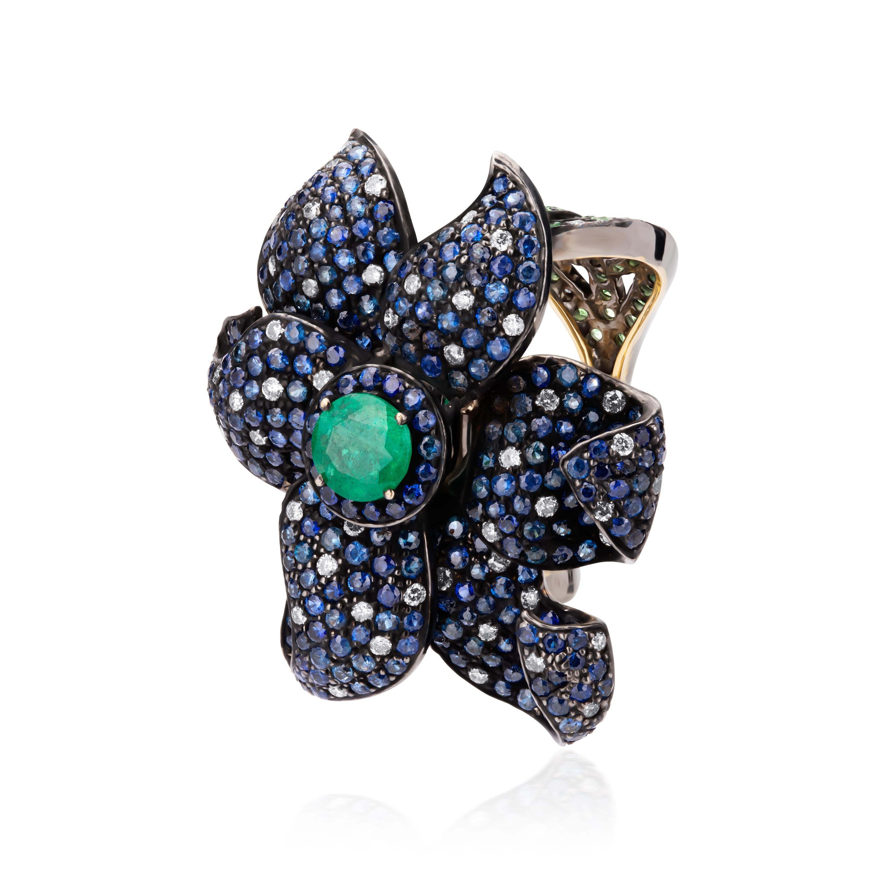 A classic Victorian flower! This extravagant ring features a round faceted emerald at the center of a flower embedded with sparkling blue sapphires. The center flower is accentuated with two bands carved with leaf motifs embellished with tsavorites.