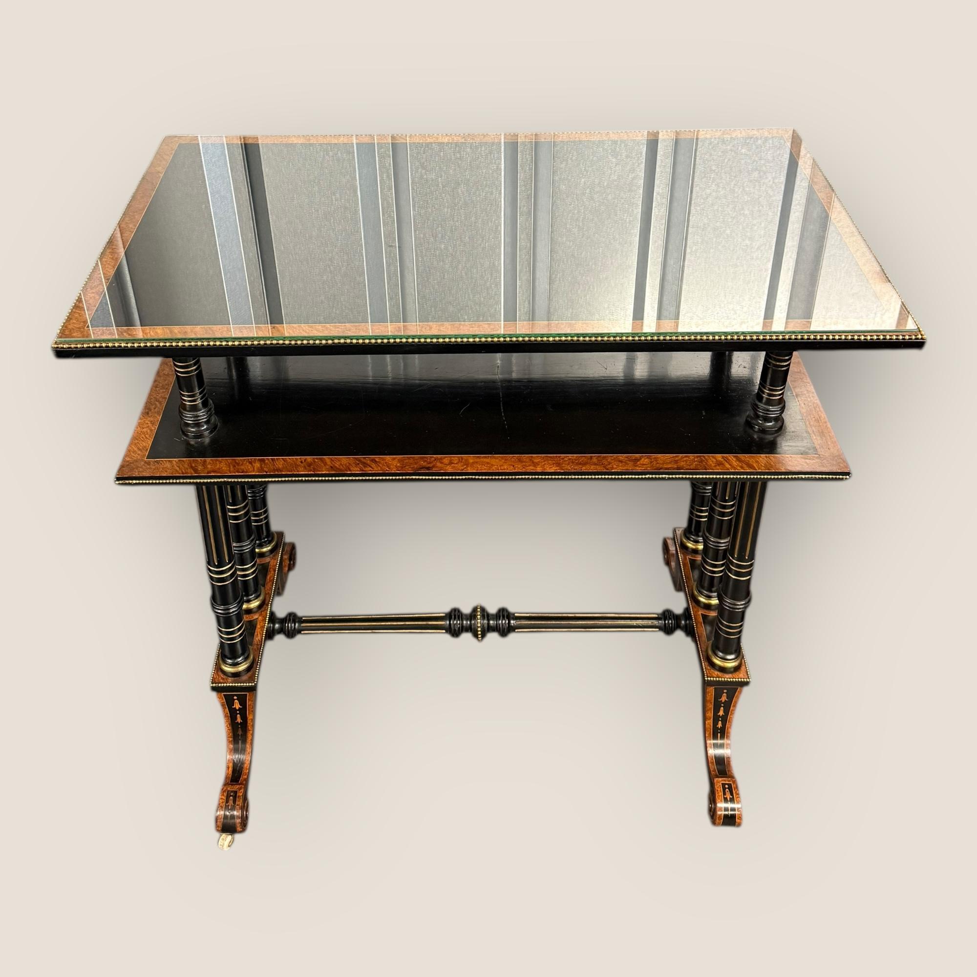 Un monte-plats victorien du mouvement esthétique en ronce de noyer et en ébène. Deux niveaux, marquetés, pieds dorés et bordés de laiton de haut en bas. Le tout est complété par un plateau en verre. Avec des roulettes en porcelaine à la base de