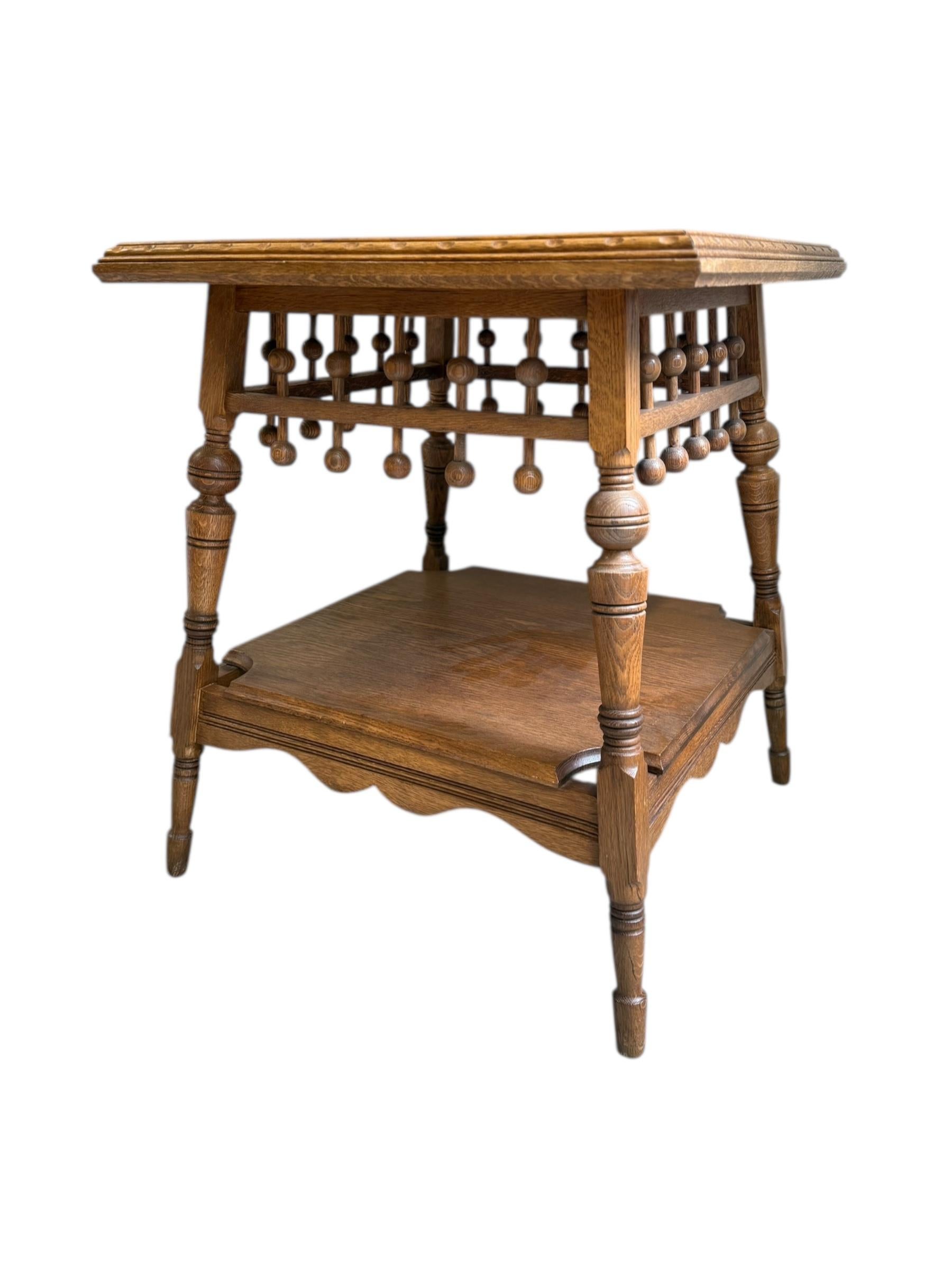 The table is a small, square, two-tiered wooden piece with a rich oak finish. It features intricately carved legs with turned spindles and bulbous details, supporting a scalloped lower shelf and a flat top surface with a molded edge. The upper tier