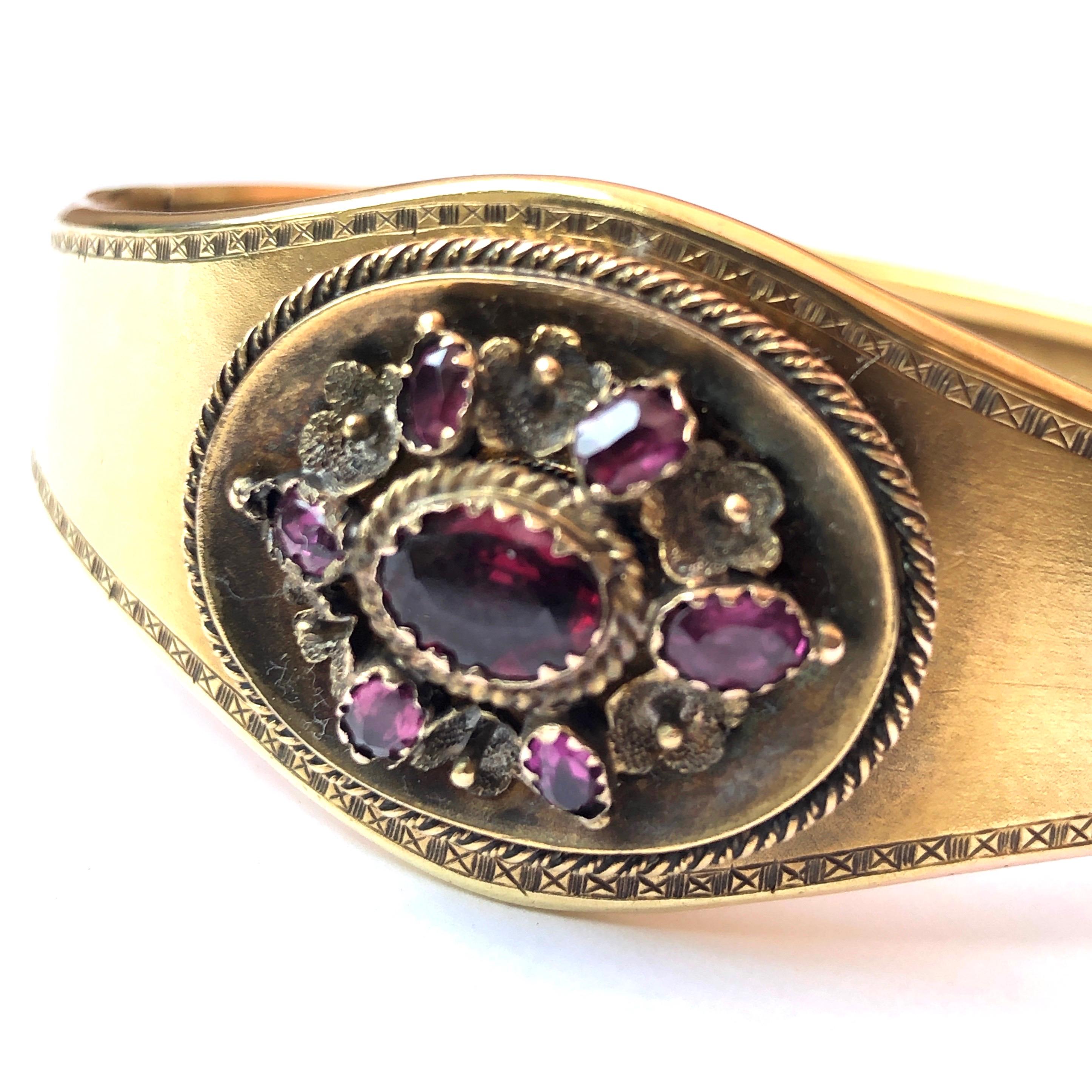 This classic style bangle holds a panel with an almandine garnet cluster and gold rope twist detail. The central stone is larger and surrounding it are six smaller stones. At the back of this panel there is a glazed locket. Modelled in 18ct gold.